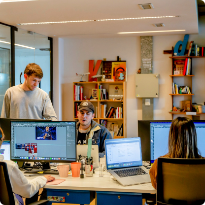 Personas trabajando en una oficina moderna con computadoras, estanterías, y una pizarra blanca.