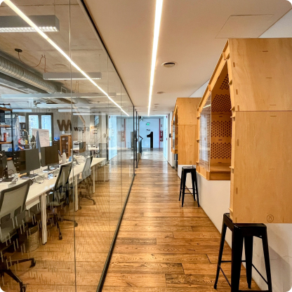 Oficina moderna con paredes de vidrio, escritorios con computadoras y sillas, suelos de madera, y decoración de madera en el pasillo.