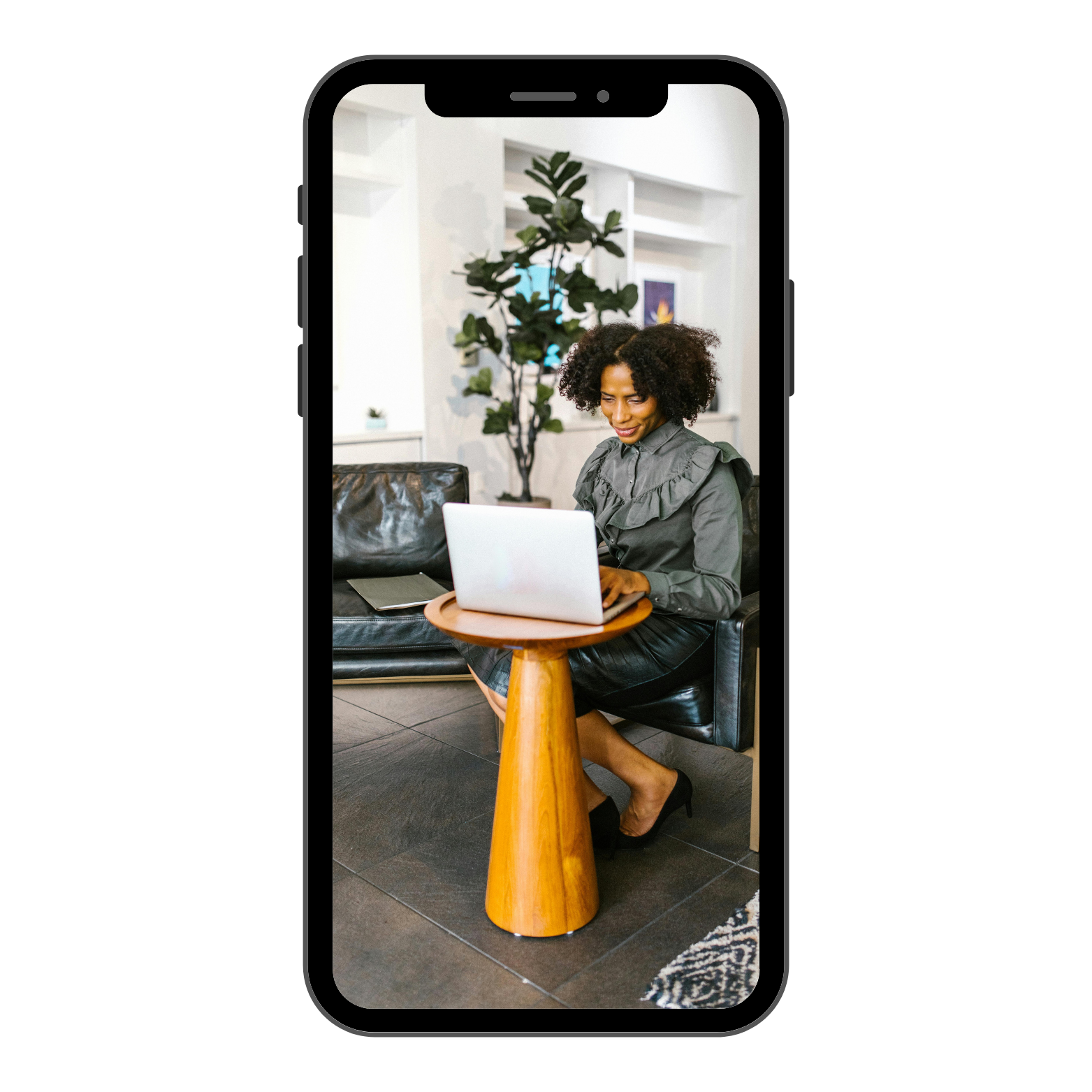 A woman with curly hair in a gray blouse and black skirt, sitting on a black sofa, working on a silver laptop at a small round wooden table in a modern, well-lit living room with a large potted plant behind her.