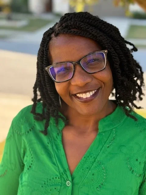 A woman with glasses and braided hair smiling outdoors, wearing a green blouse.
