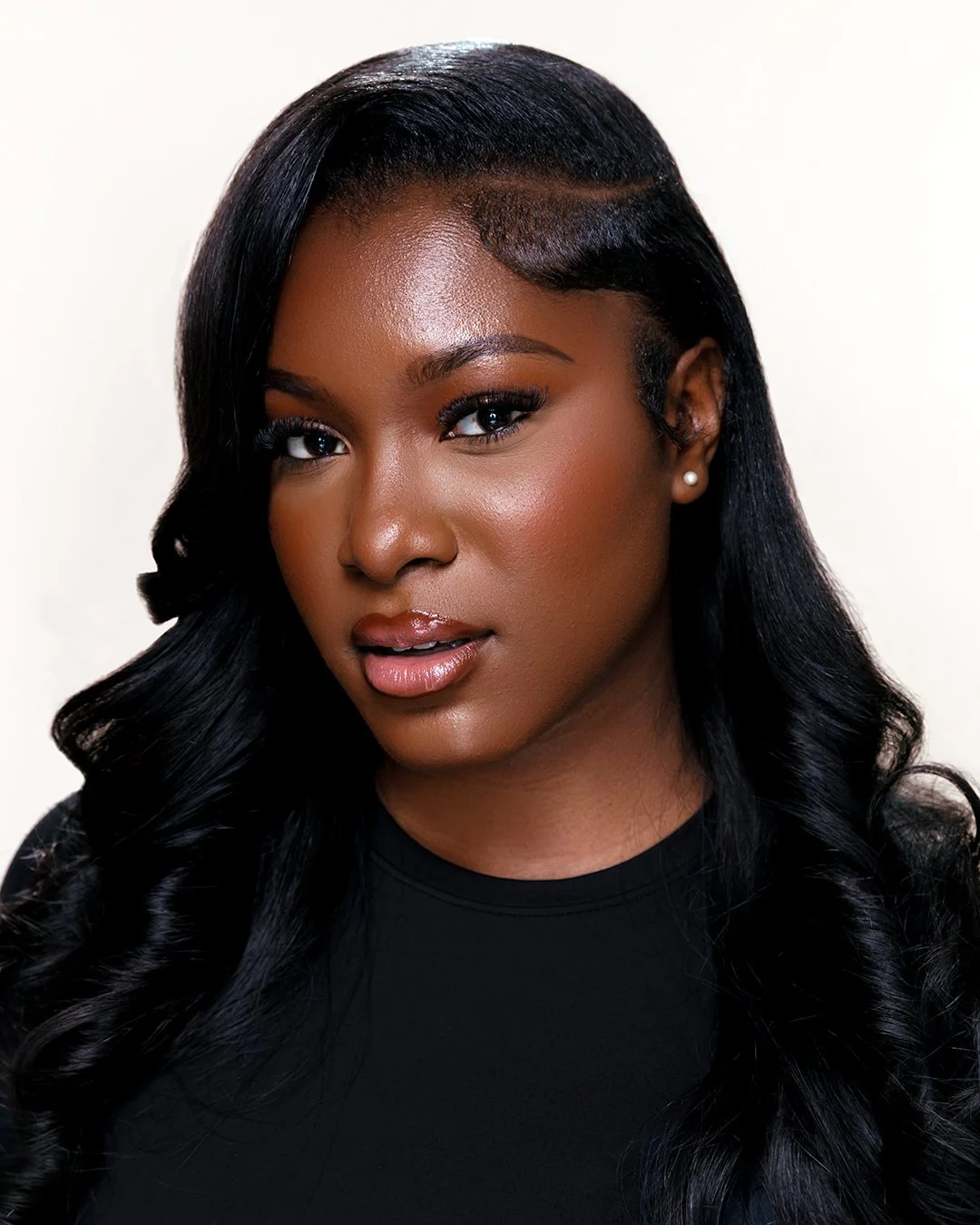 A close-up portrait of a young Black woman with long, wavy black hair, wearing pearl earrings and makeup.