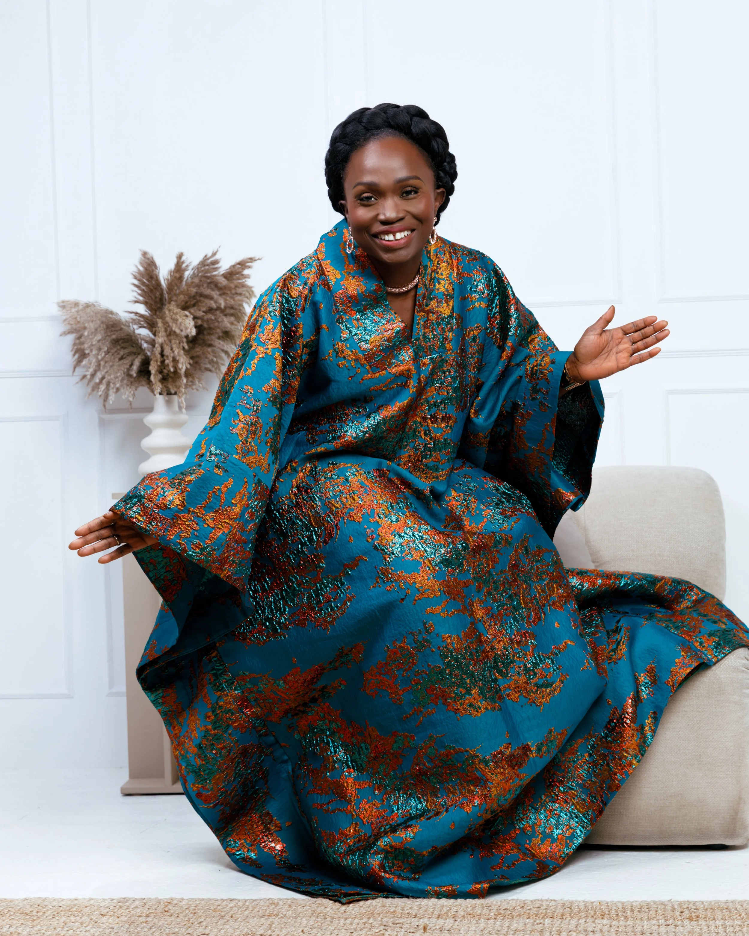 A woman wearing a colorful, shiny blue and orange patterned dress, smiling and sitting on a beige sofa, with a white wall and a tall vase of pampas grass in the background.