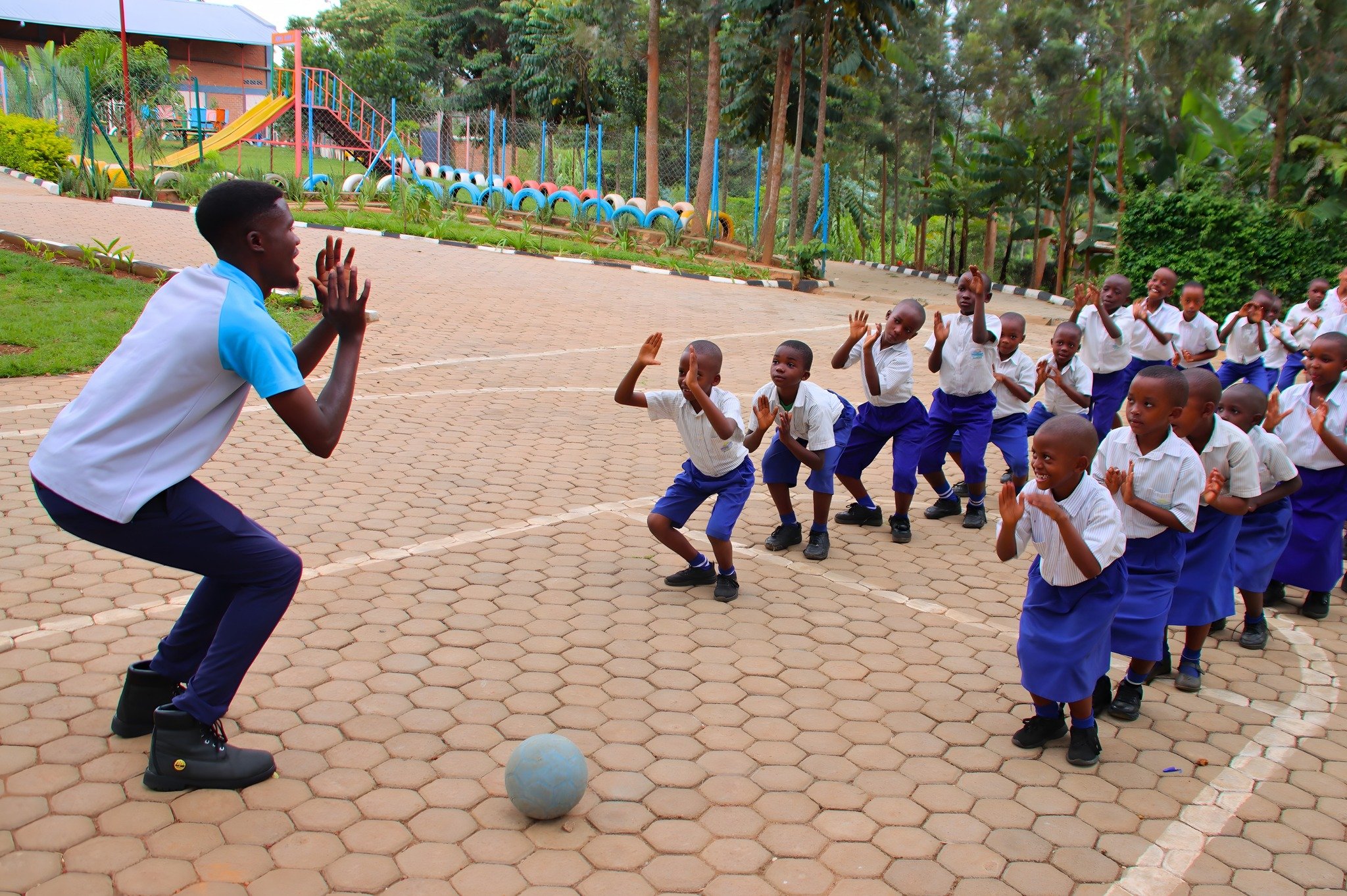 Aristophane Igiraneza, our outstanding Physical Education (PE) teacher at RCCS, is instructing a group of Primary students on the fundamentals of volleyball. Although the pose Aristophane and the children have struck in the picture may seem unusual, 