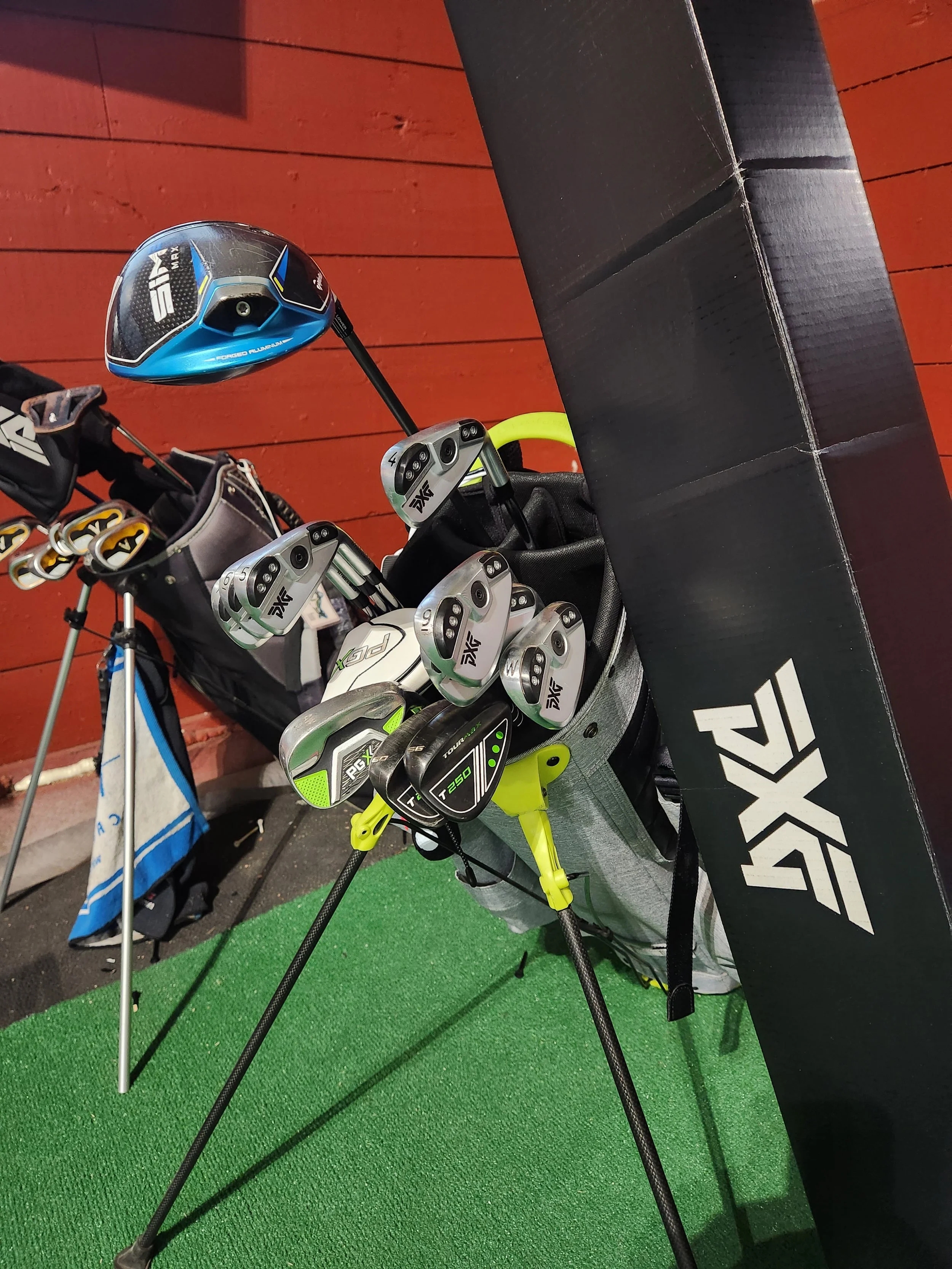 Set of golf clubs on a stand, featuring a mix of PXG and SIM Max brands, against a red wall background on a green carpet.