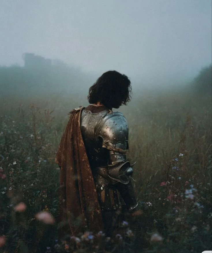 A person dressed in medieval armor standing in a foggy field with wildflowers, their head bowed and face partially hidden.
