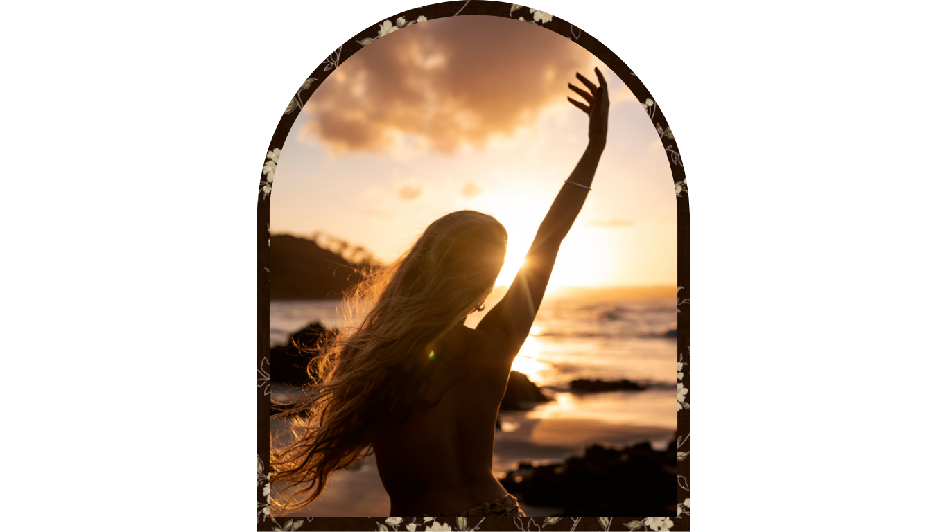 A woman with long hair standing on a beach during sunset, with her arm raised towards the sky.