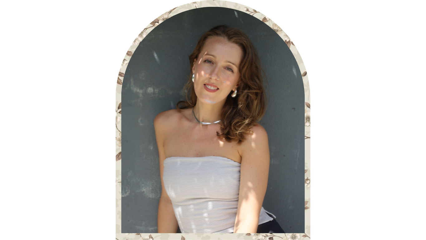 A woman with shoulder-length curly brown hair, wearing a strapless white top, silver earrings, and a silver choker, smiling while leaning against a gray wall.