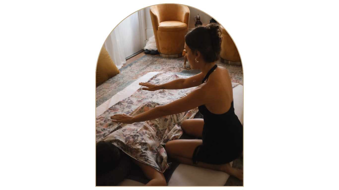 A woman practicing yoga or stretching on a bed in a cozy, well-lit room with patterned carpet, pillows, and vintage-style chairs.