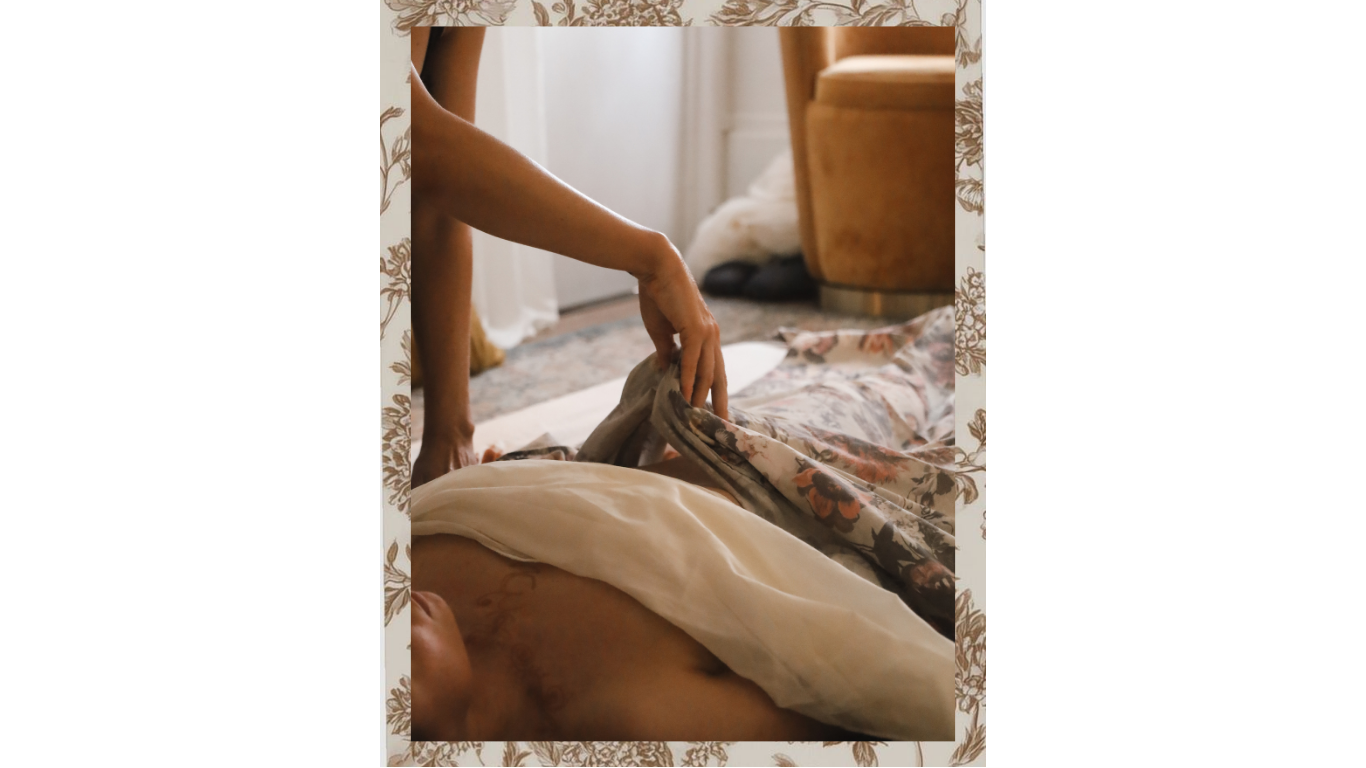 Person lying on bed with floral sheets, another person adjusting the sheets, in a cozy room with a brown armchair and a window.