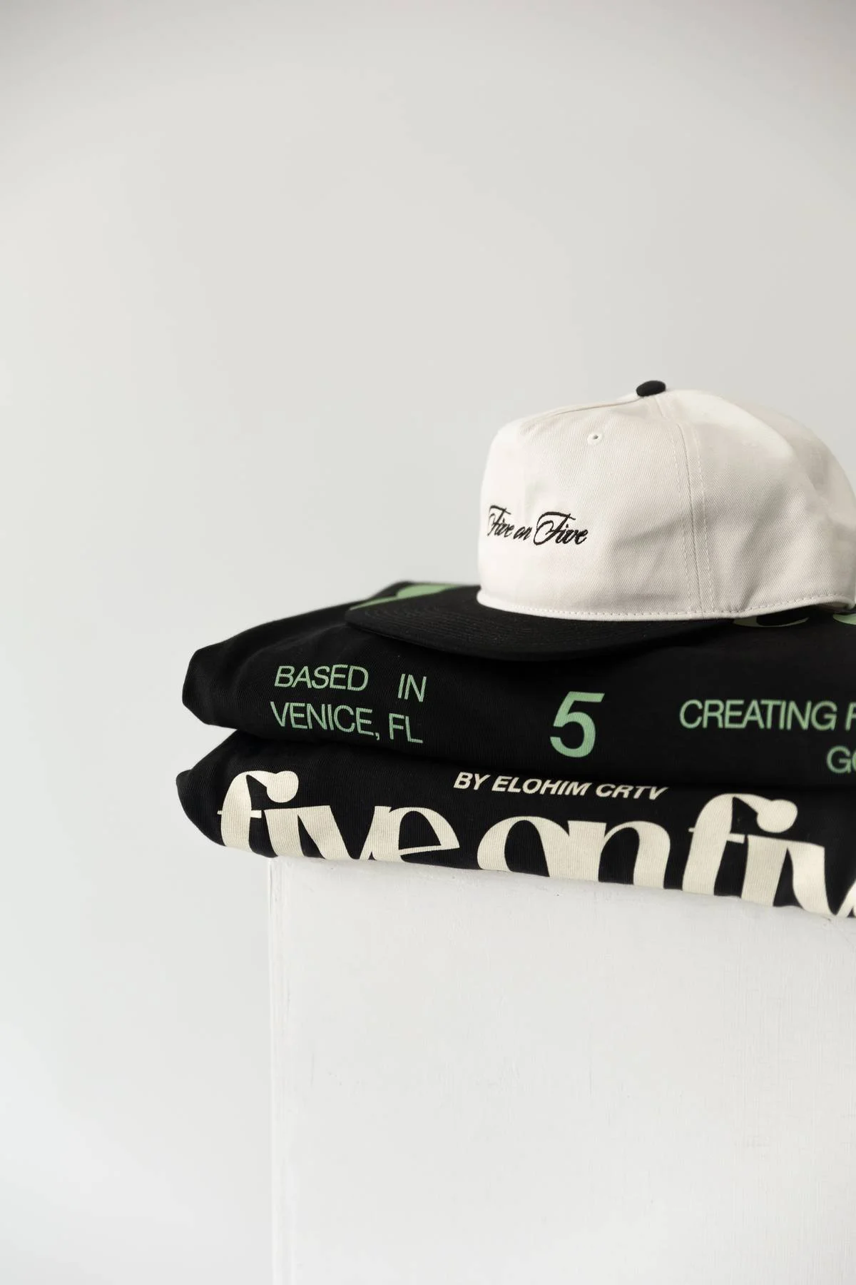 A white baseball cap with black embroidery reading 'Fear en Fivé' on top of two folded black t-shirts with green and white text.