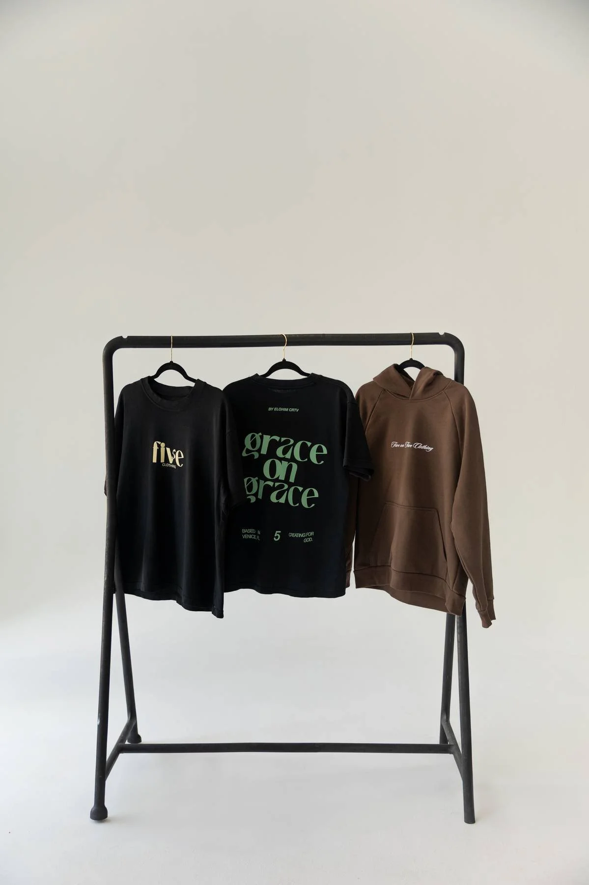 Three casual clothing items hanging on a black clothing rack against a plain white wall. From left to right: a black t-shirt with the word 'five' in gold, a black t-shirt with green text 'grace on grace', and a brown hoodie with white embroidered tex