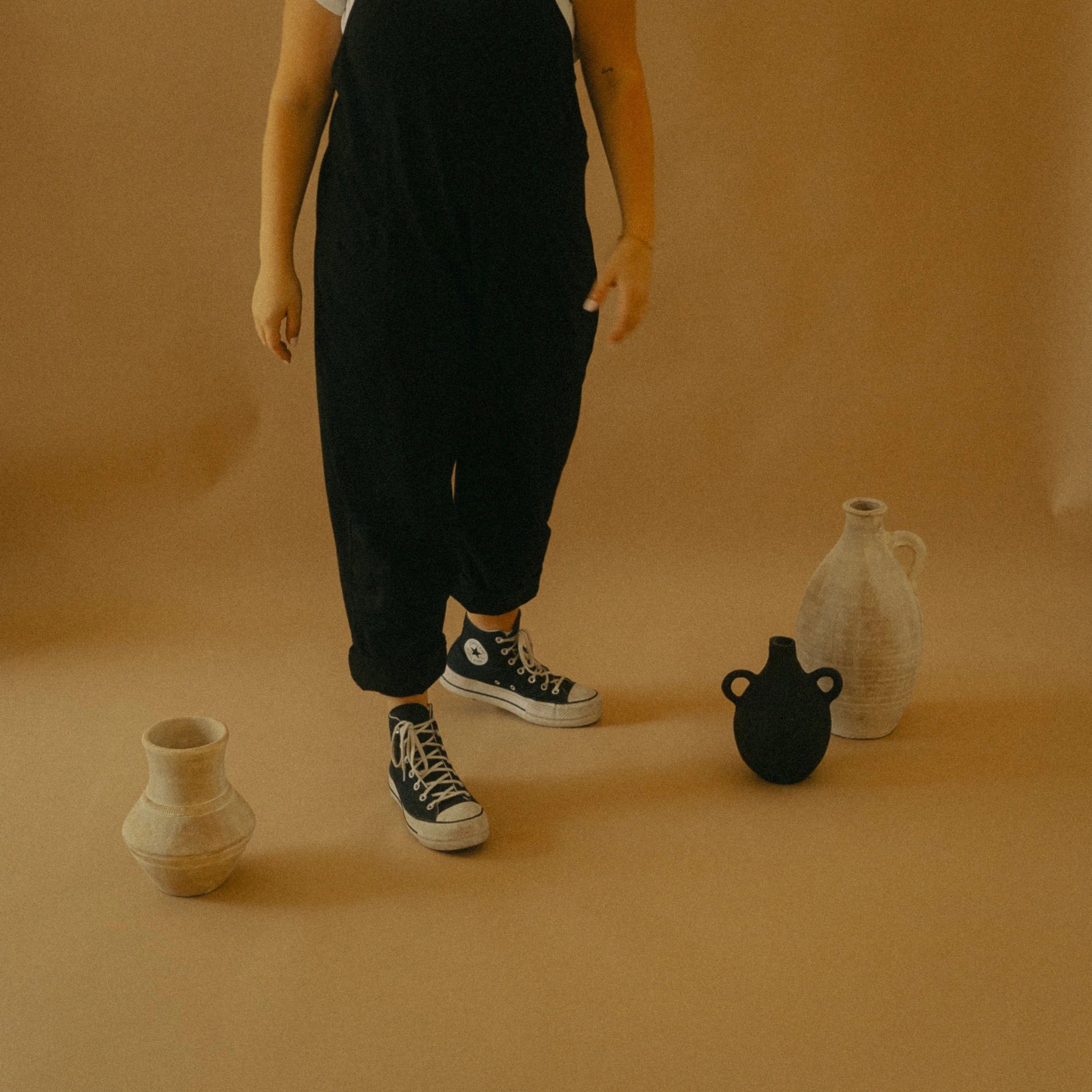 Person wearing black jumpsuit and sneakers standing next to three vases on beige background.