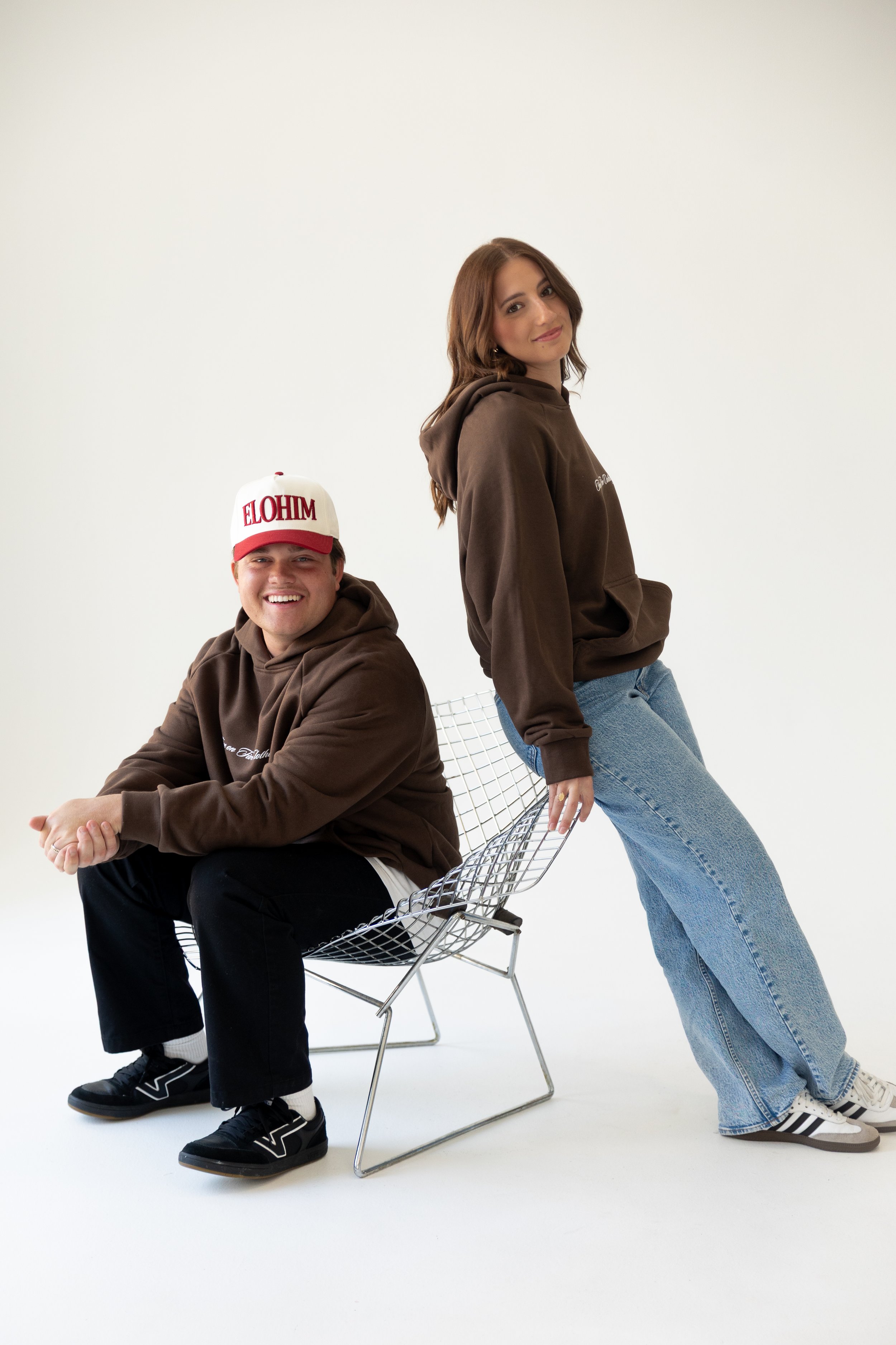 A young man and woman in brown hoodies, with the man sitting on a wire chair and the woman leaning against it, smiling and posing against a white background.