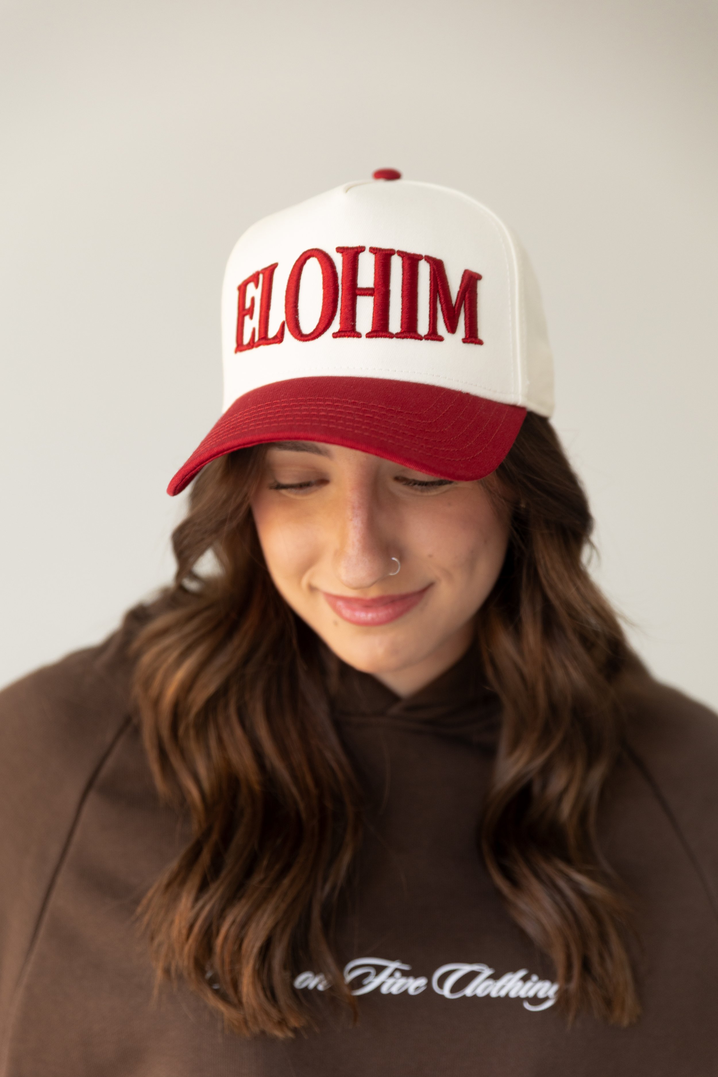 A woman with long wavy brown hair, wearing a brown hoodie with white text, and a white and red trucker hat with the word 'ELOHIM' embroidered on the front, smiling with her eyes closed.