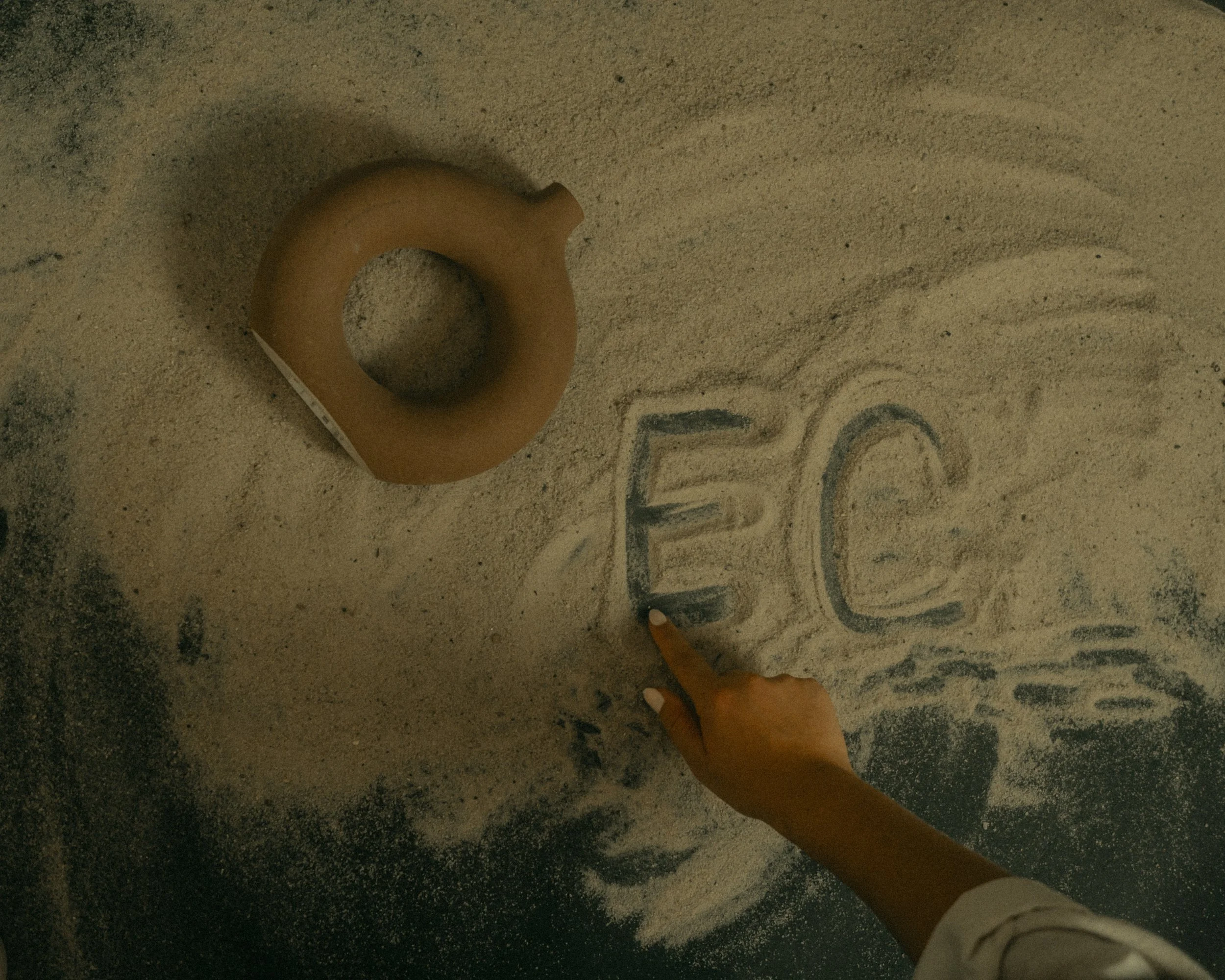 A hand writing the letters 'E' and 'C' in blue chalk on a sandy surface, with a wooden ring-shaped object nearby.