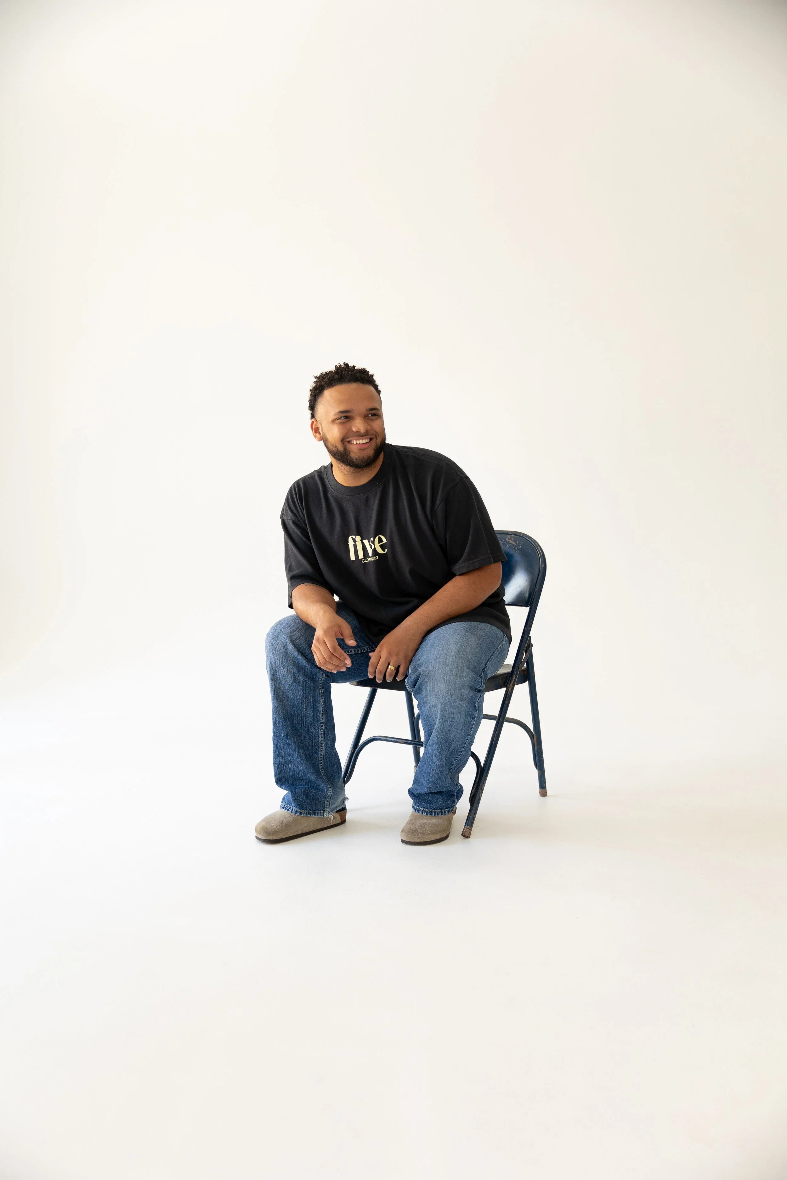 A smiling man with a beard and curly hair sitting on a blue folding chair in a white studio, wearing a black T-shirt with the word 'five' printed on it, blue jeans, and gray shoes.