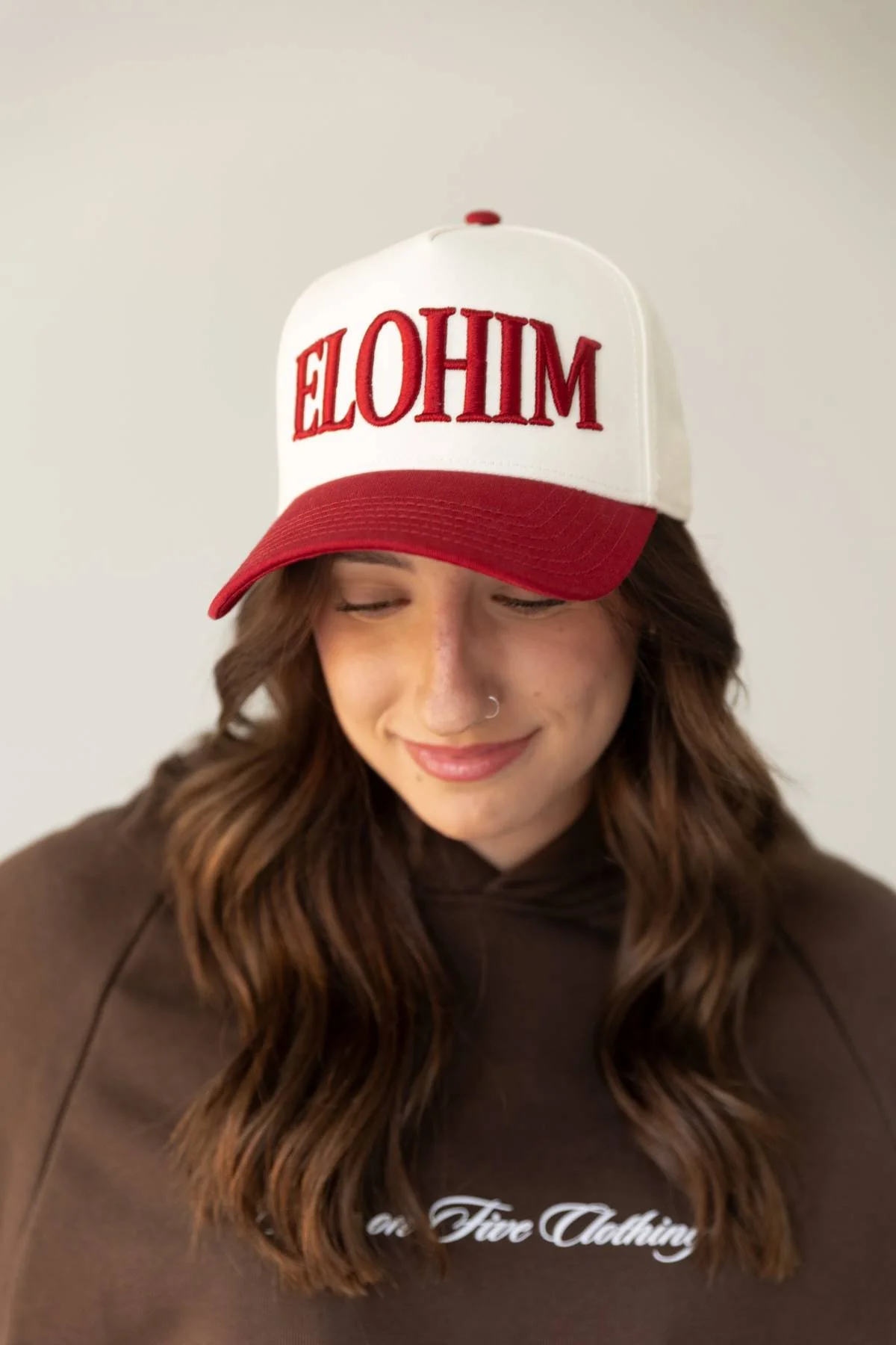A woman with wavy brown hair and a nose piercing is wearing a red and white trucker hat with upside-down red embroidered text, and a brown hoodie with white cursive text.