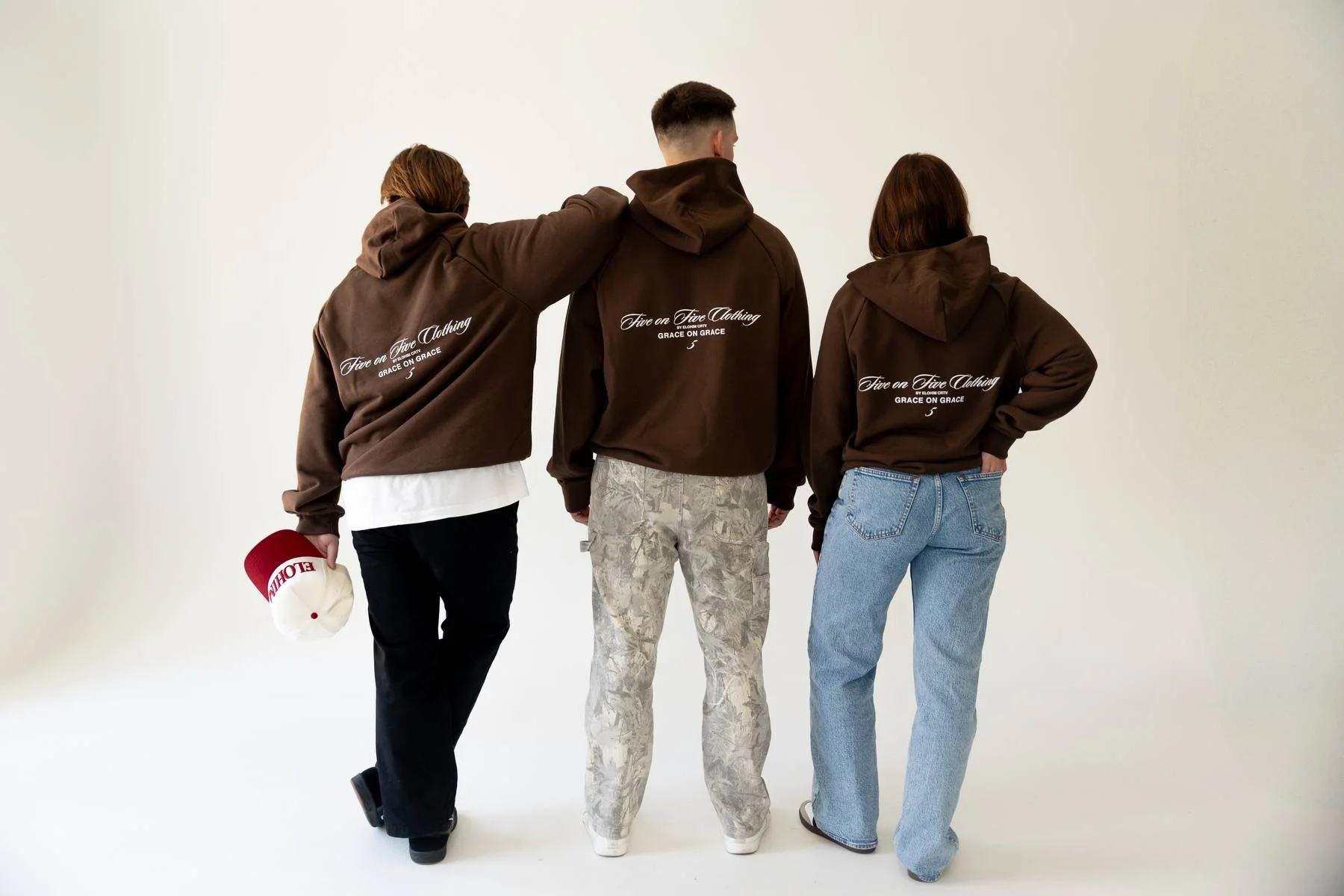 Three people in brown hoodies with white text on the back, standing with their backs to the camera; one person is holding a red and white cap.