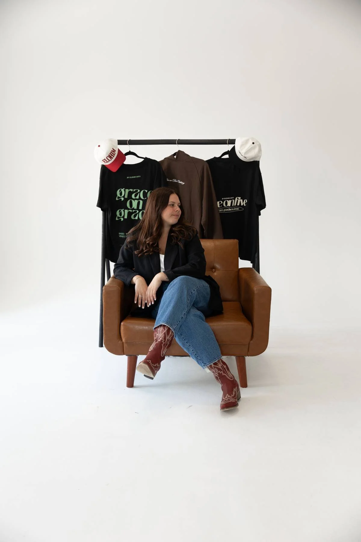 A woman sitting on a brown leather armchair with a clothing rack behind her. The rack has three hats and three shirts with text on them. The background is plain white.