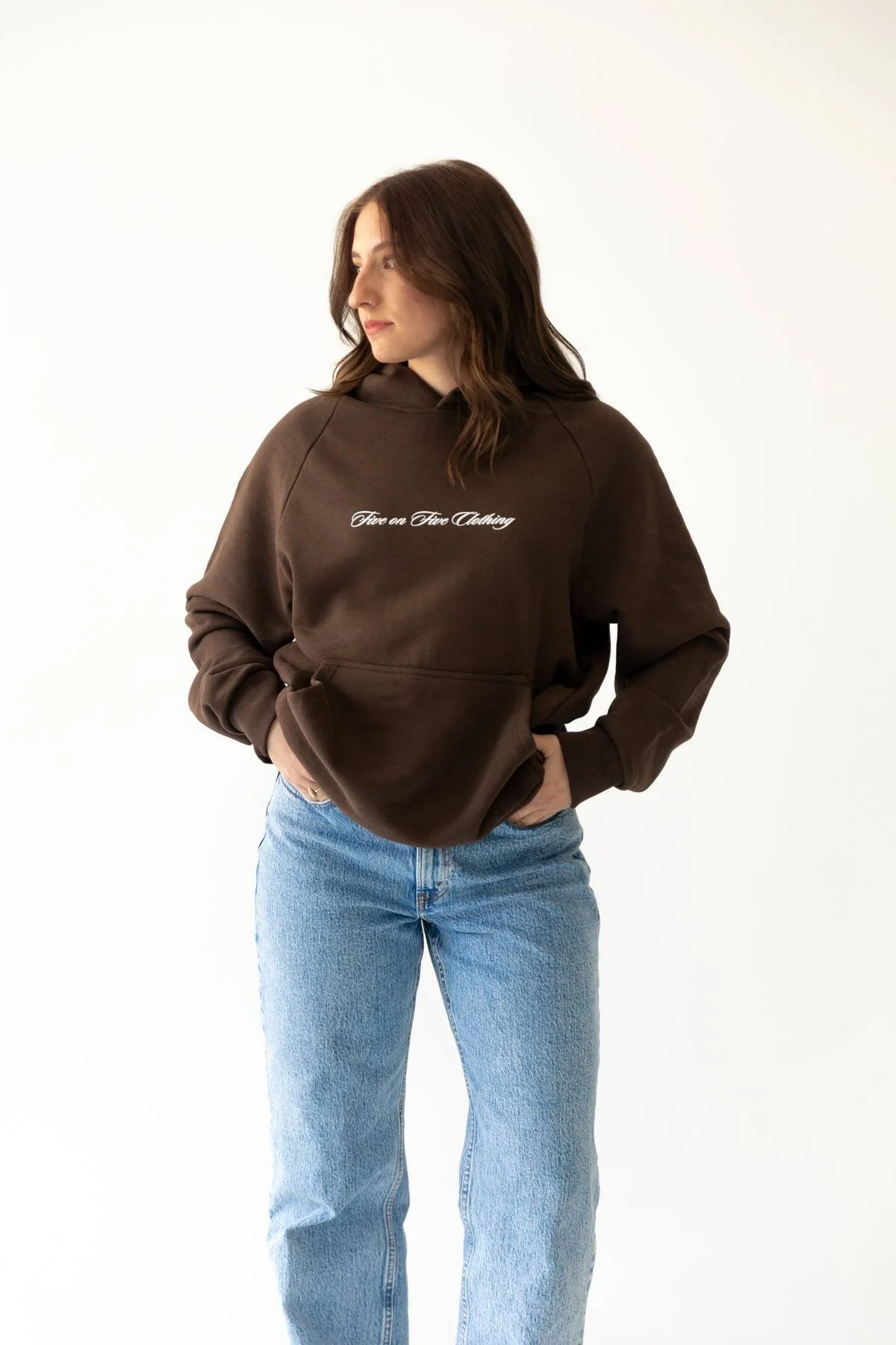 A woman with brown hair wearing a dark brown hoodie with white text and light blue jeans standing against a plain white background.