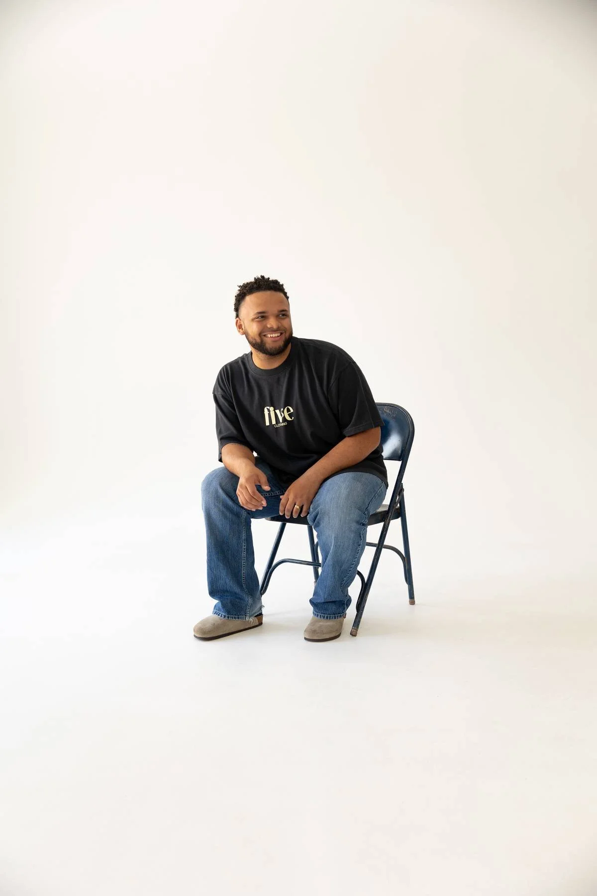 A man with dark curly hair and a beard sitting on a metal folding chair in a white studio background, smiling, wearing a black T-shirt with the word 'five' printed on it, blue jeans, and beige shoes.
