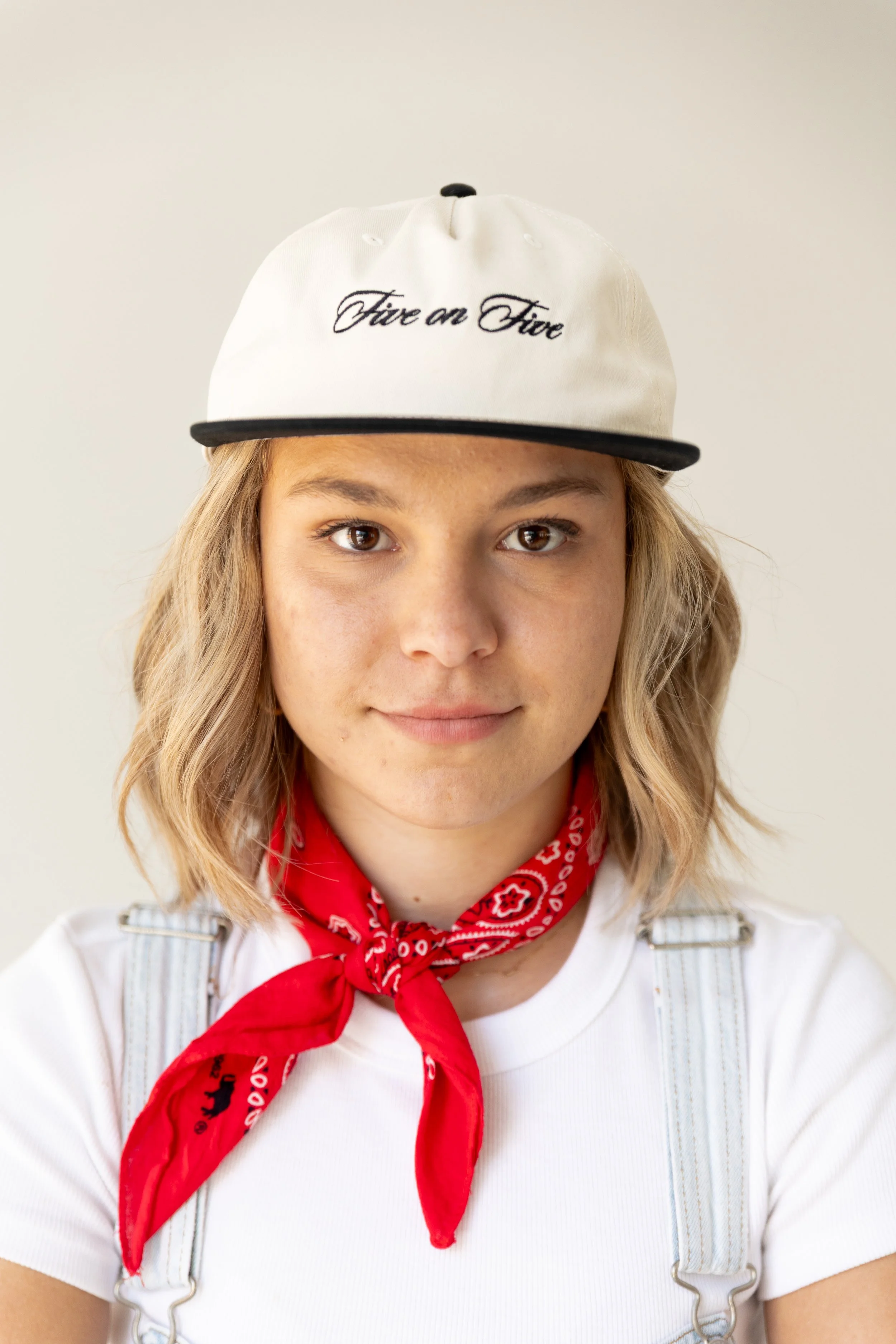 A young woman wearing a white baseball cap with black embroidery that says 'Fire and Fire,' a white t-shirt, a red bandana around her neck, and denim overalls, looking directly at the camera with a subtle smile.