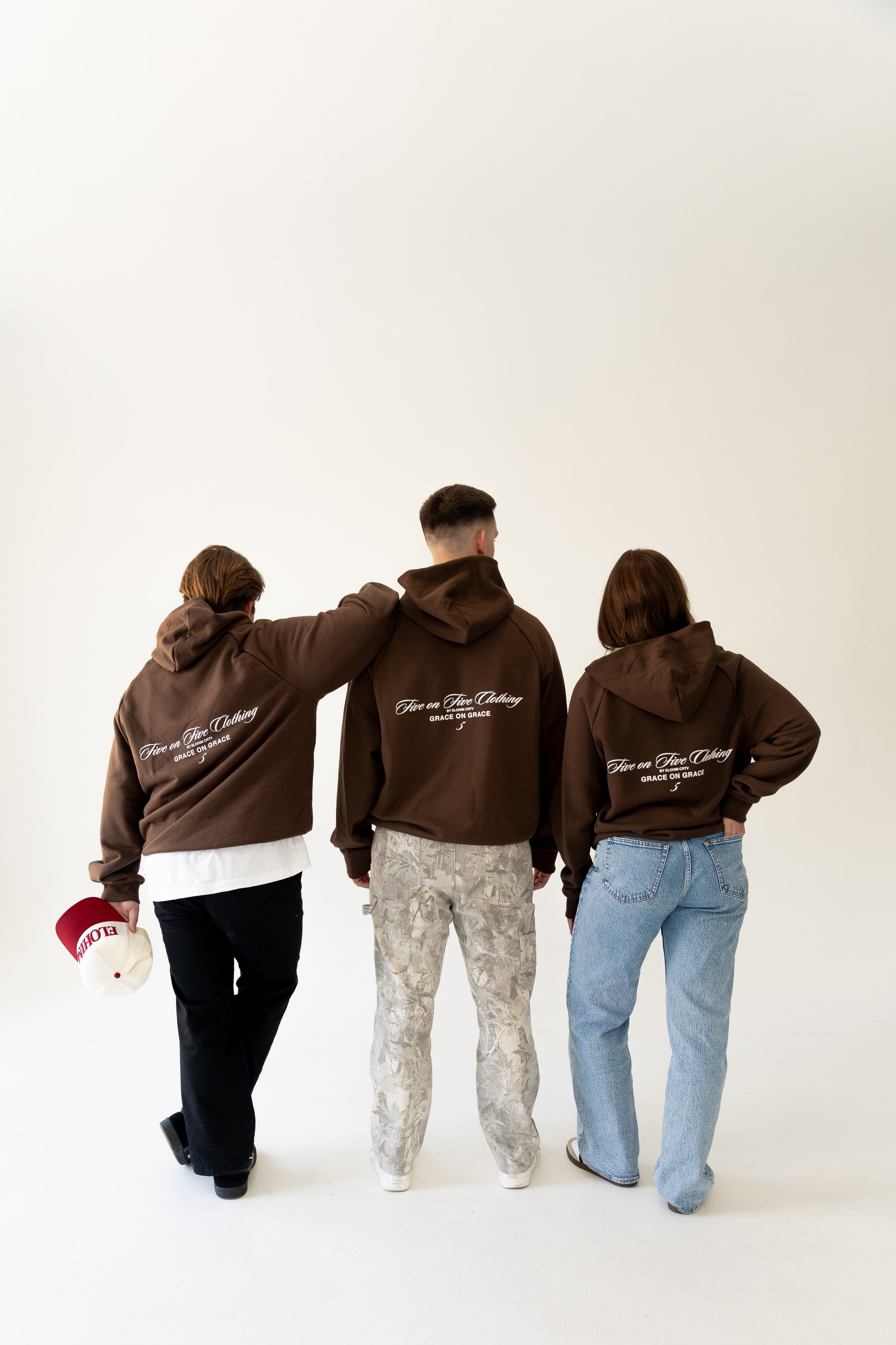 Three people from behind wearing matching brown hoodies with text, standing against a plain white background. The person on the left holds a red and white cap.