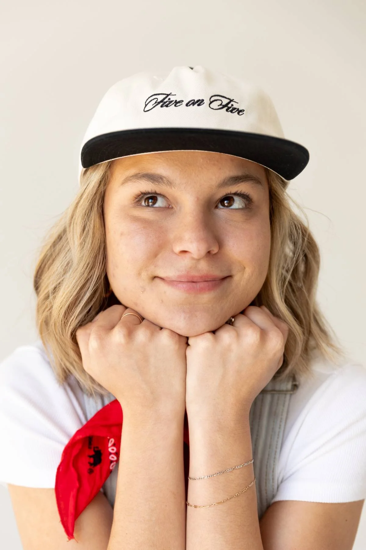 A young woman with blonde hair, wearing a white cap with black brim that says 'Fire on Fire', resting her chin on her hands, smiling softly, with a red bandana around her neck and jewelry on her arms.