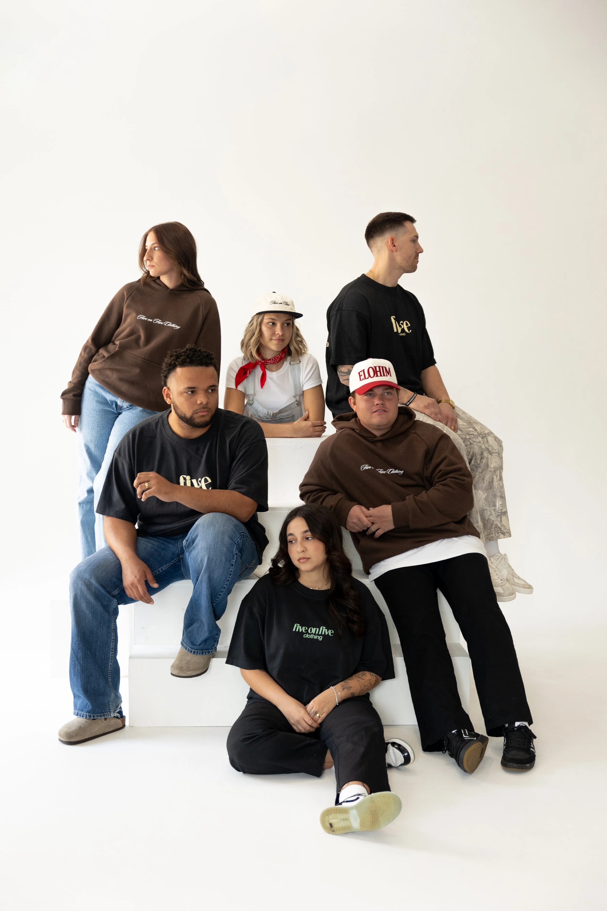 Group of six young adults posing on a white background, wearing casual streetwear including hoodies, t-shirts, and caps.