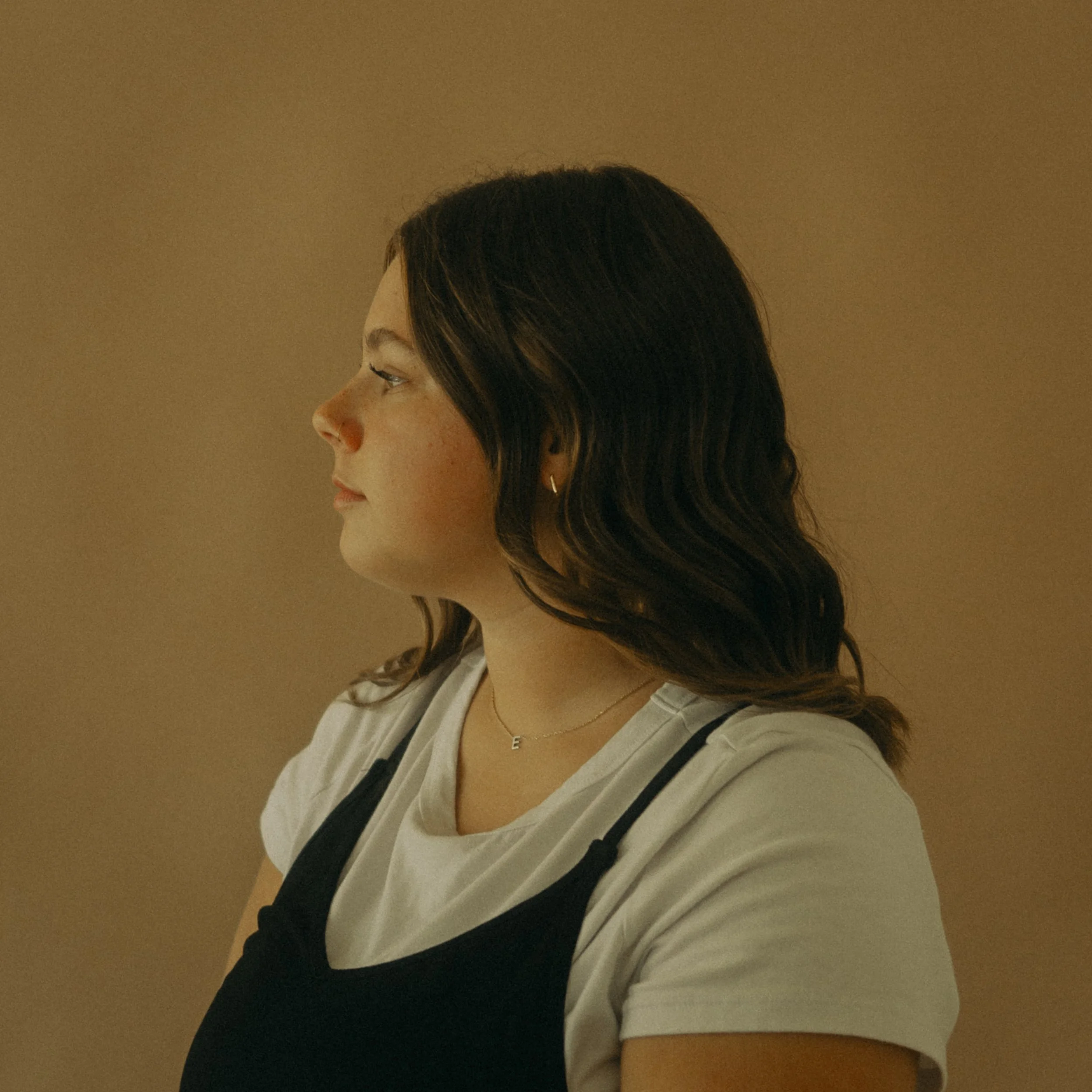 Side view of a woman with wavy brown hair wearing a white t-shirt, a black top, a small letter 'E' necklace, and small hoop earrings, standing against a beige background.