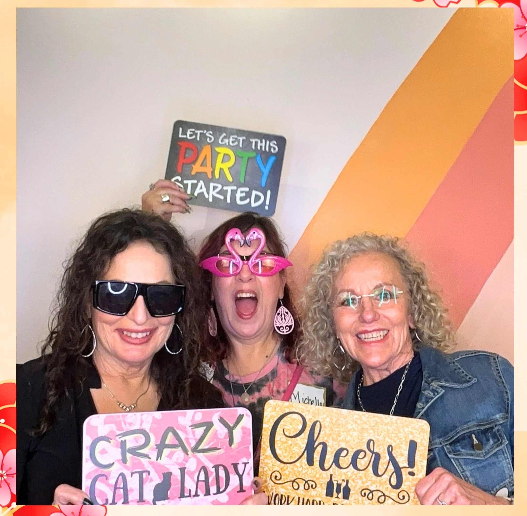 A group of women at a celebration holding colorful signs with words like "celebrating" and "cheers". They are wearing glasses, earrings, and pink party accessories, smiling and enjoying the moment.