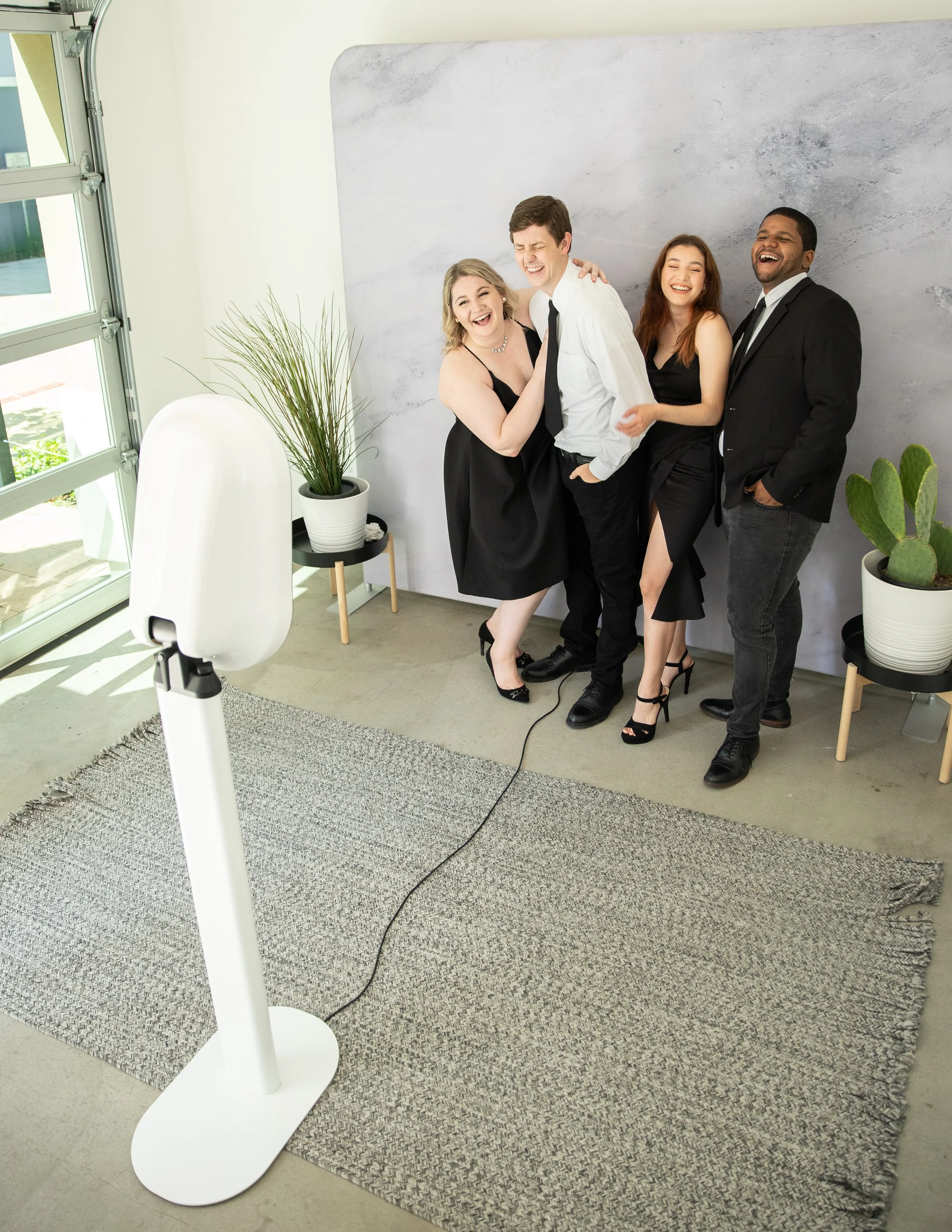 Five people in formal attire laughing and smiling in front of a gray marbled wall, with two potted plants and a white chair nearby, at a photo shoot setup.