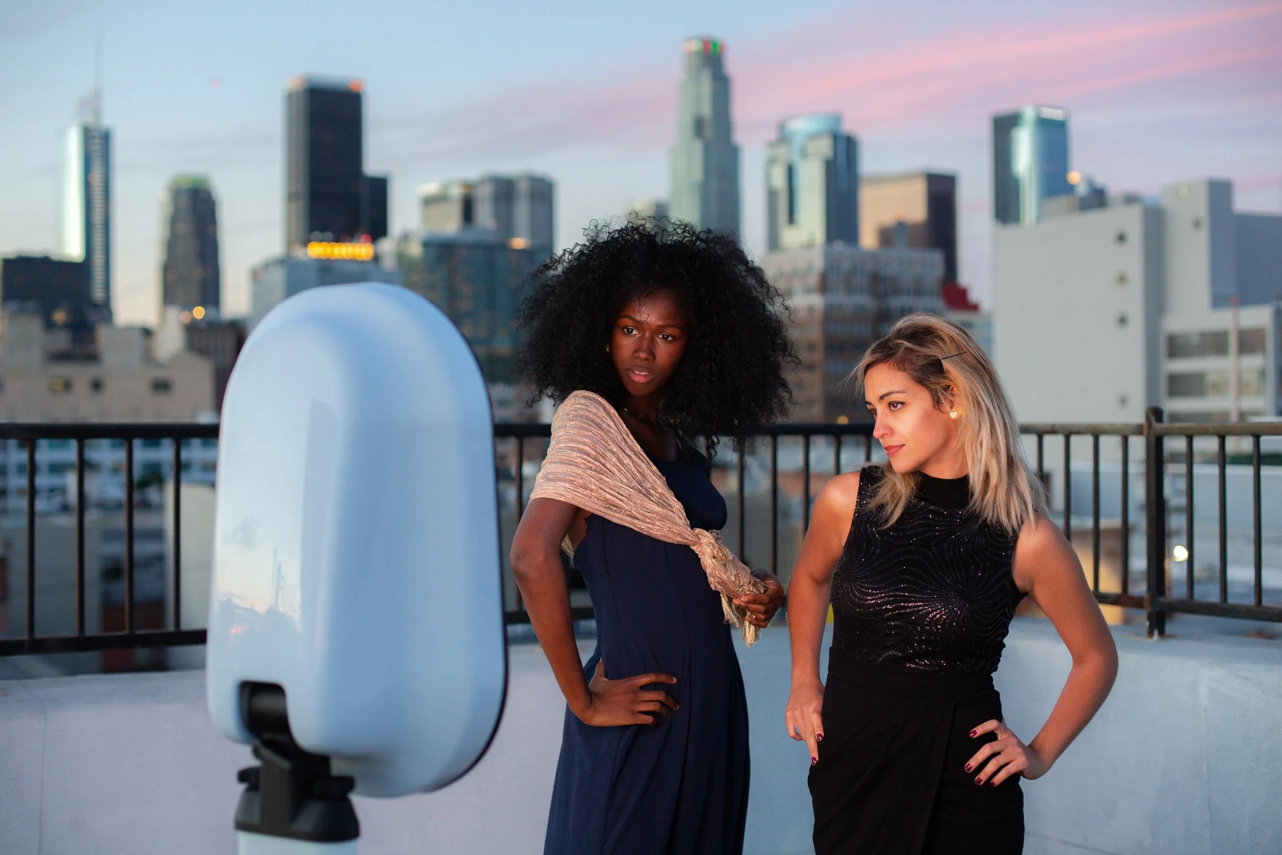 Two women stand on a rooftop at sunset, posing in front of a photobooth with a city skyline in the background.