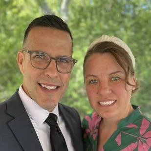 A man in a suit and tie with glasses and a woman with a headband smiling outdoors against a green, leafy background.