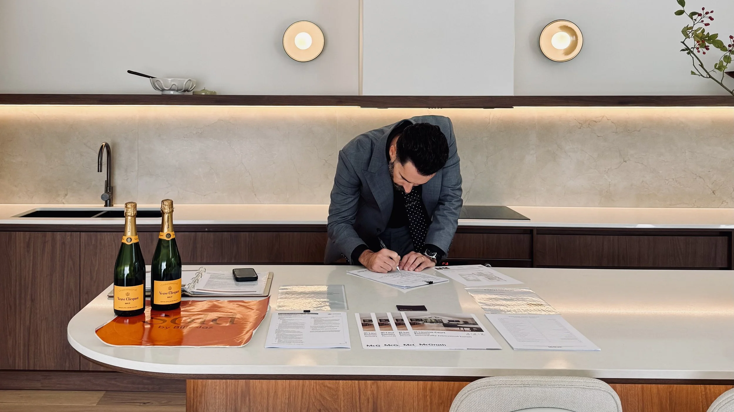 A man in a gray blazer is signing documents on a white kitchen counter with papers, a phone, and two bottles of Veuve Clicquot champagne placed on an orange cloth. Billy Jaz McGrath Estate Agent Auctioneer Palm Beach Currumbin Waters Palm Beach