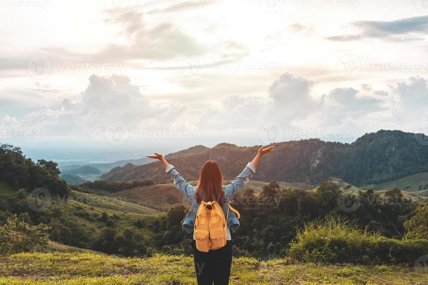 A woman with a yellow backpack standing on a grassy hill with outstretched arms, overlooking a scenic mountain landscape under a cloudy sky during daytime.