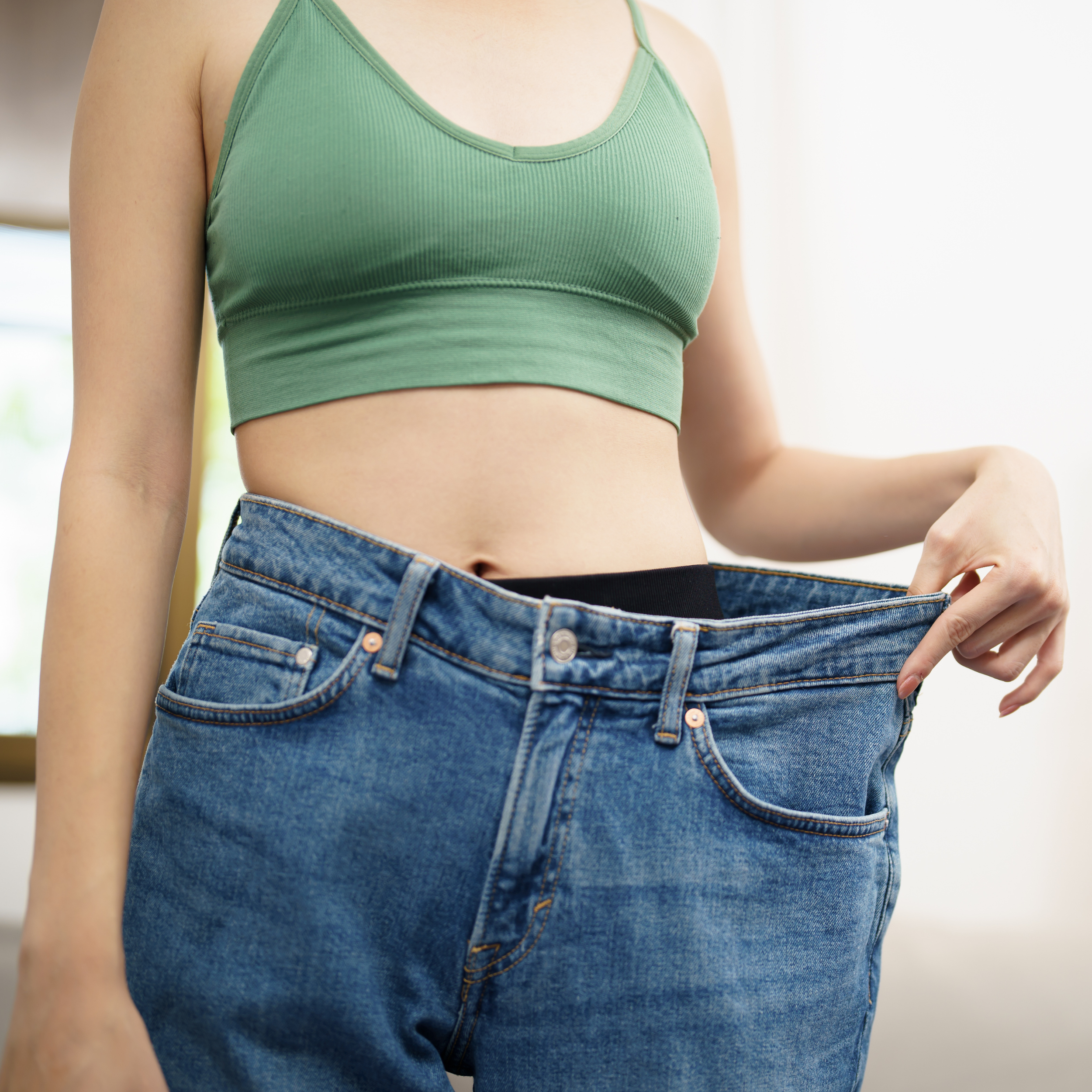 Person wearing a green sports bra and oversized blue jeans, holding out the waistband to show it's too loose.