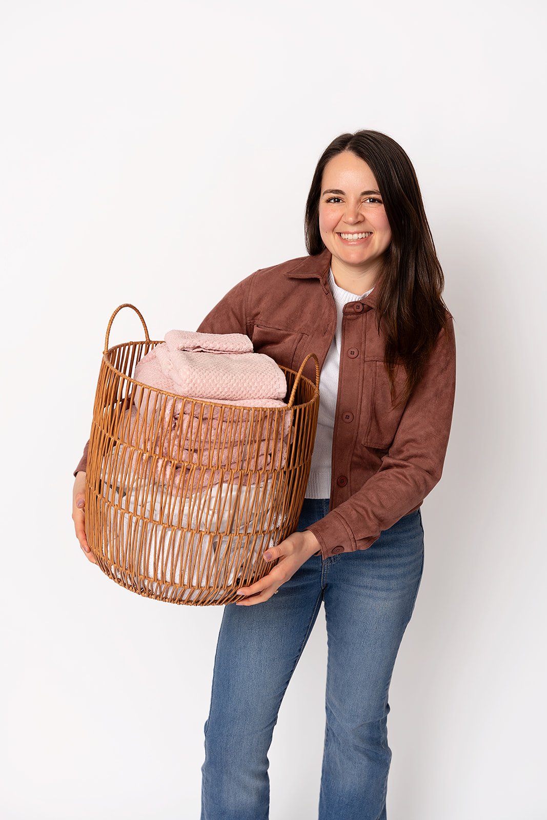 A woman with brown hair smiles at the camera while holding an organized wicker basked of pink linens. She is wearing blue jeans, a white shirt, and a brown jacket.