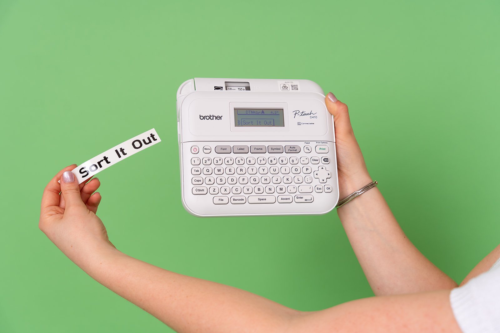 A professional organizer holds up a Brother label maker with a label that reads "Sort it Out" in front of a green background.