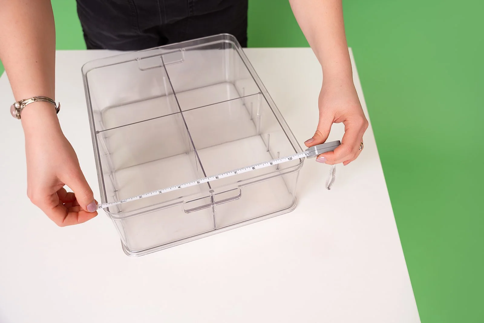 A woman holds a measuring tape up against a clear organizing tray that sits on a white table with a green background.