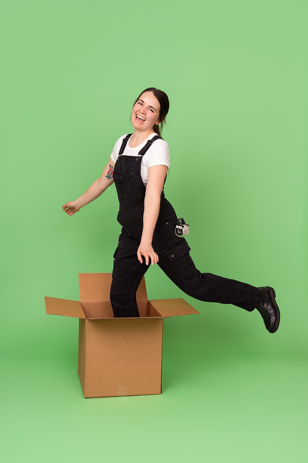 A woman with brown hair stands in a cardboard box smiling in front of a green background. She is wearing a white tee shirt and black overalls and has a tape dispenser on her hip.