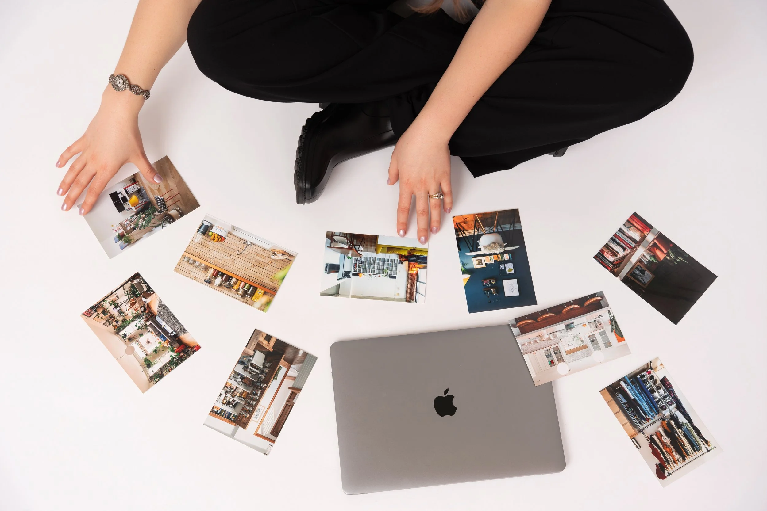 A woman sits on the floor in black pants with images of organized homes spread out on the floor in front of her next to her laptop.