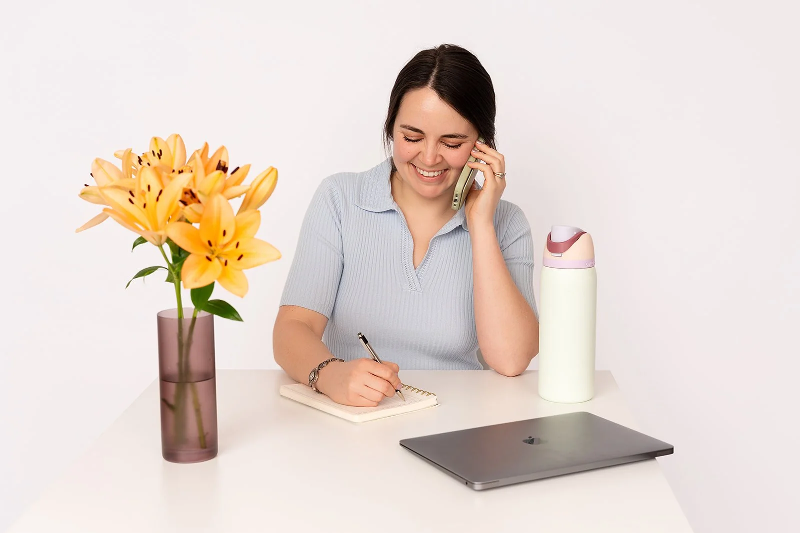A woman sits at a table with yellow flowers, a laptop, and a white water bottle. She is wearing a blue shirt and writing notes while talking to someone on her cell phone.