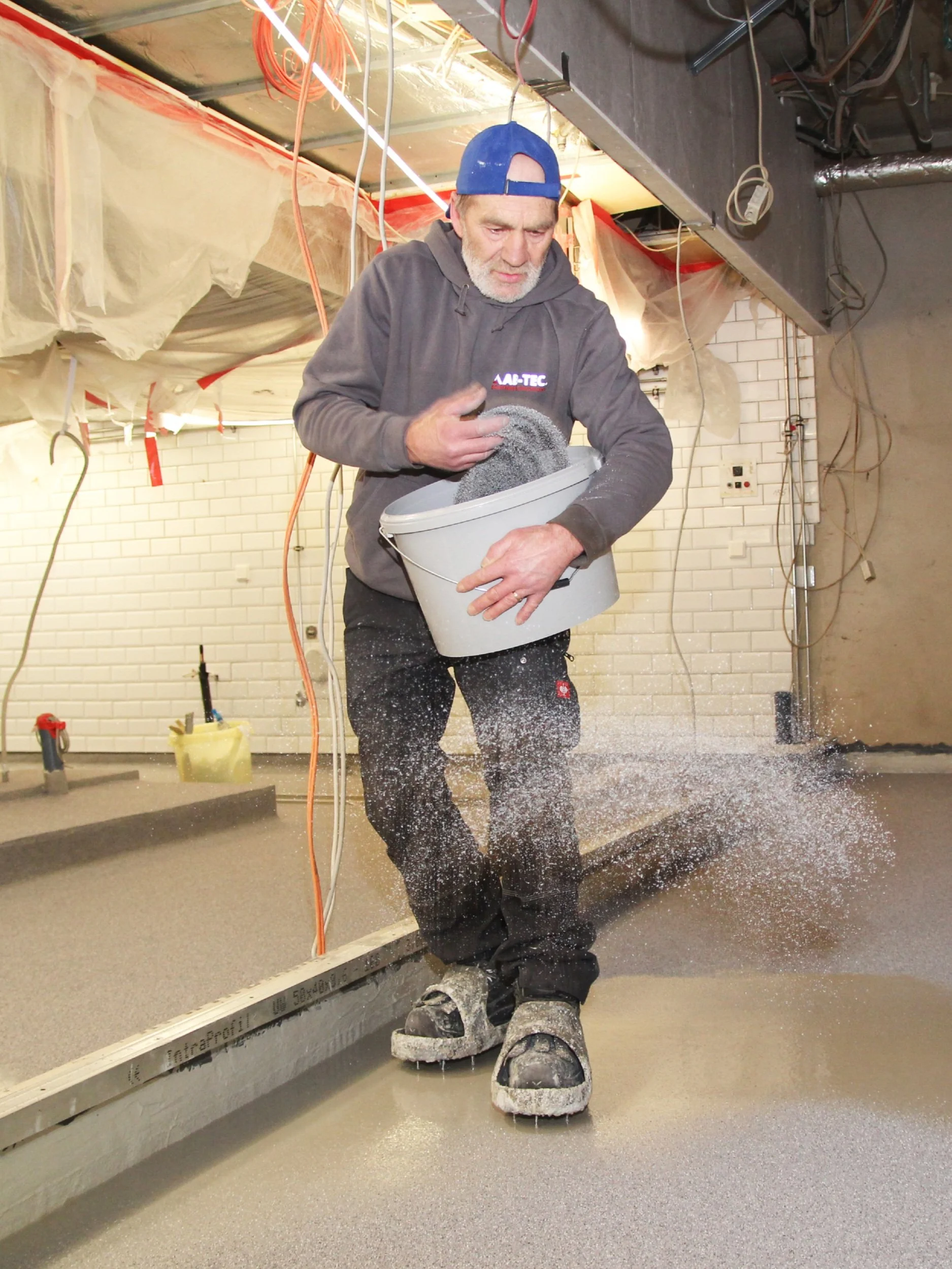 Ein Mann mit grauem Hoodie und blauer Kappe verteilt Granulat auf einem frisch verlegten Boden in einem Baustellenbereich mit freiliegenden Kabeln und Abdeckplanen.