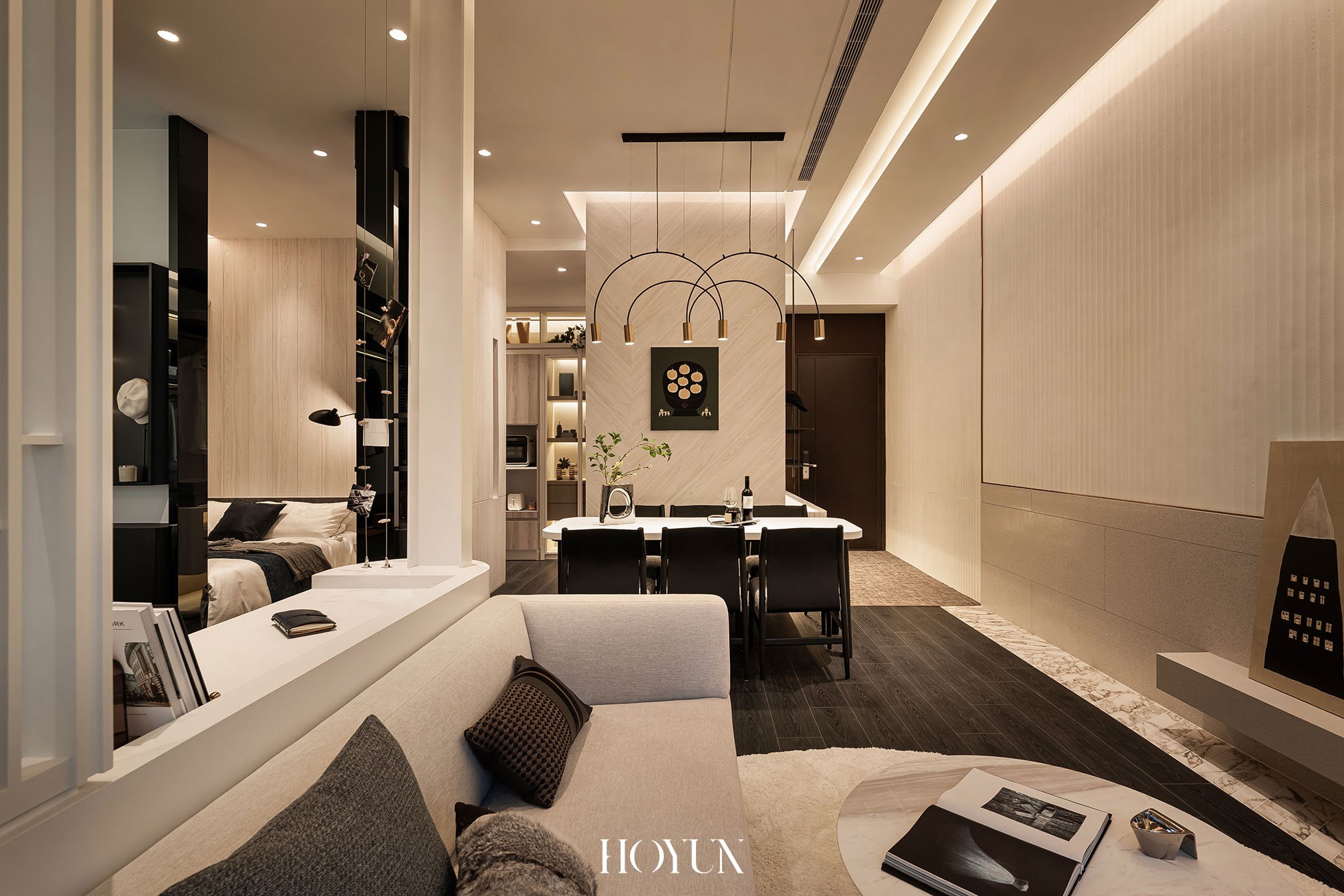 Modern living room with beige sofa, black and brown pillows, coffee table with open magazine, and a dining area with black chairs and a white table, in a well-lit open-plan space.