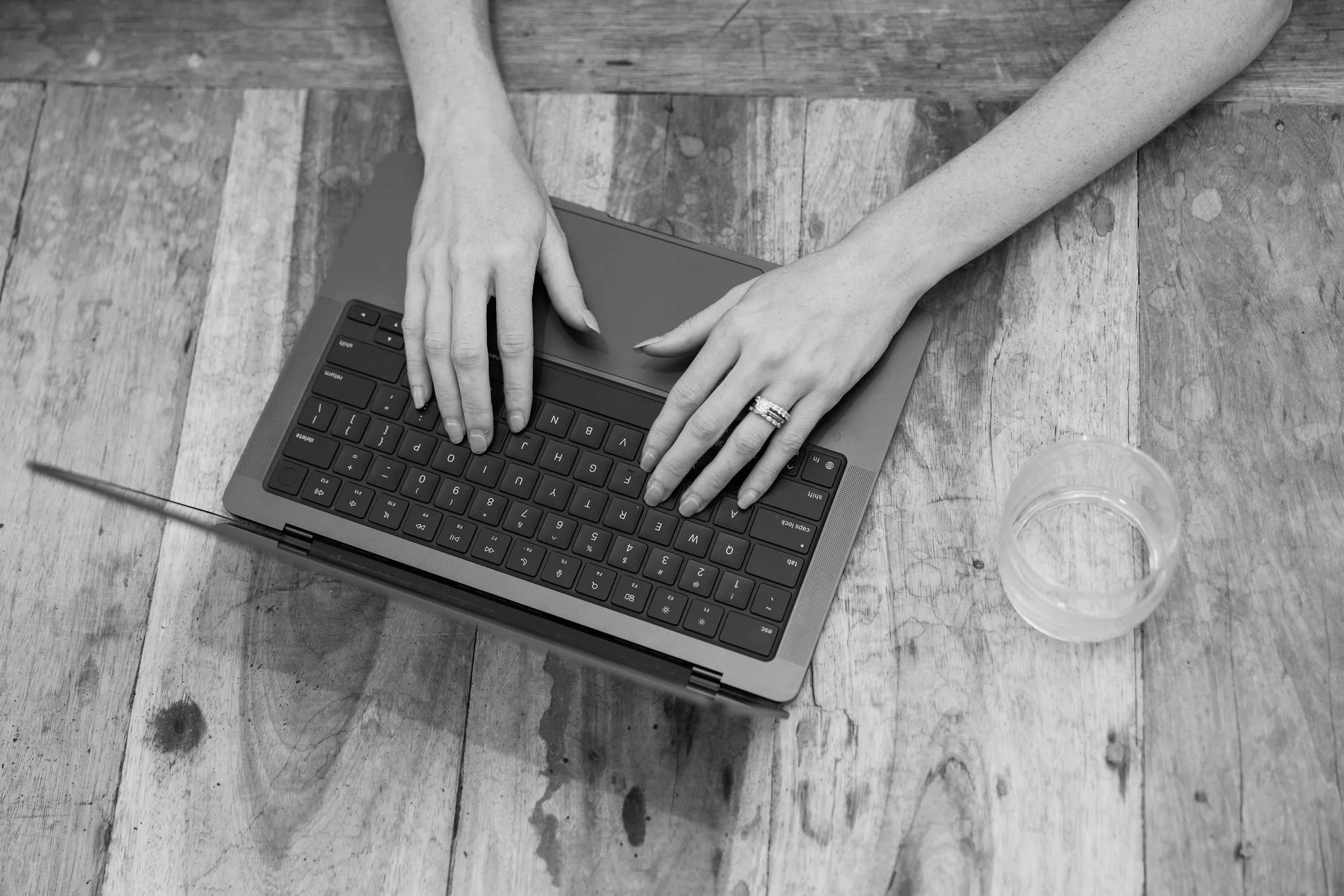Black and white image of a person's hands typing on a laptop keyboard on a wooden table, with a glass of water nearby.