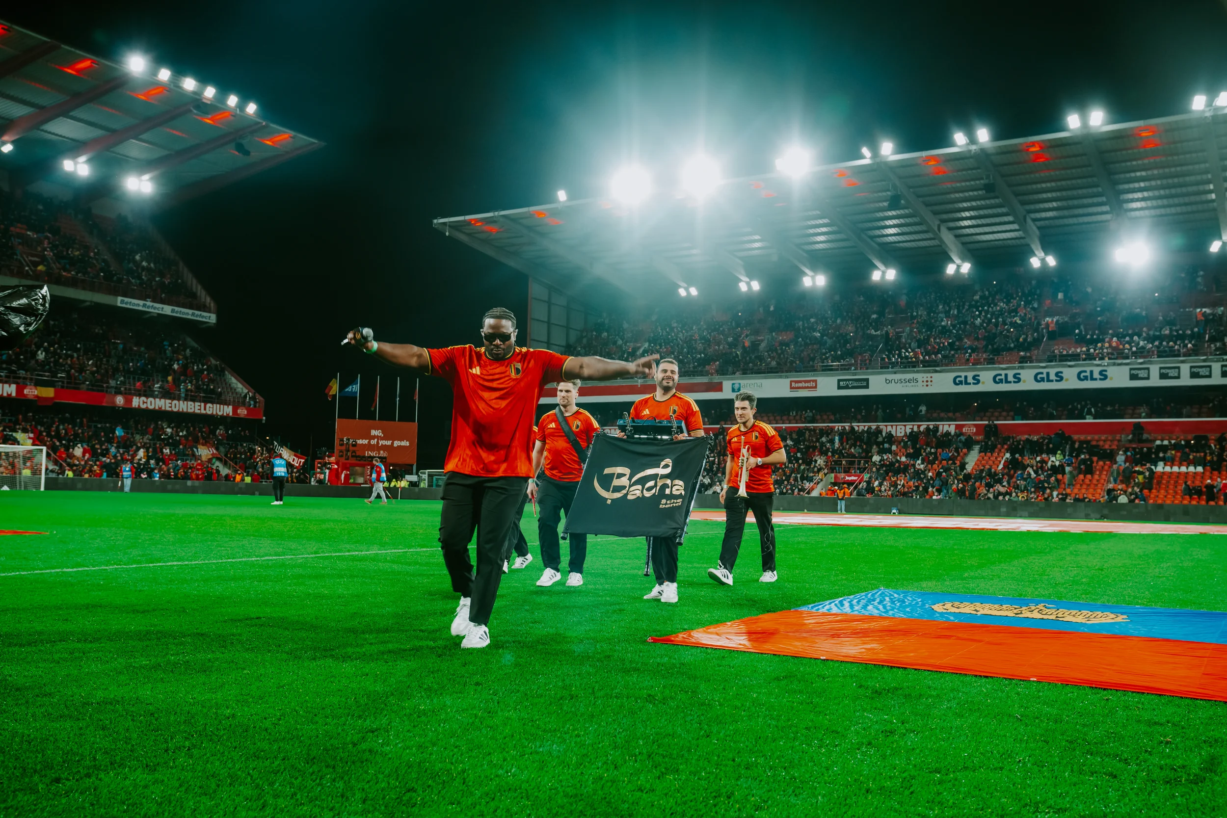 Bacha & The Band tijdens hun optreden bij de Rode Duivels. Voetbalspelers in rood ten afscheid op het veld van een stadion met een volle tribune en heldere stadionverlichting.