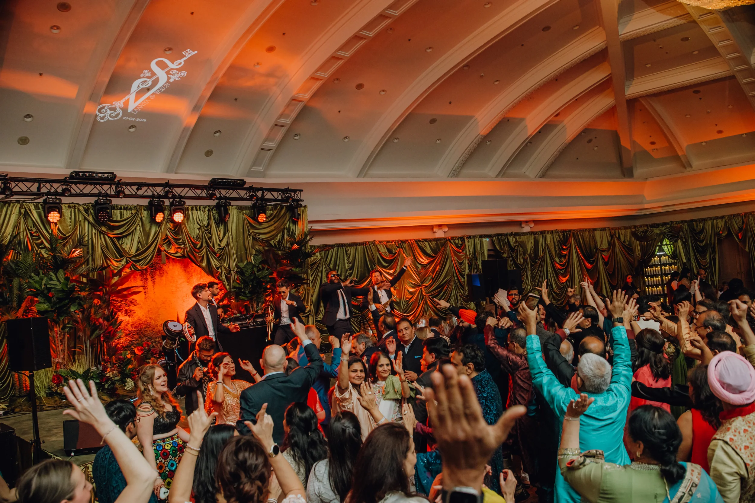 Een grote feestzaal gevuld met mensen die dansen en plezier maken. Op het podium staan muzikanten en zangers, terwijl de achtergrond versierd is met groene gordijnen en levendige decoraties. Het plafond heeft een projectie van een logo met de tekst '