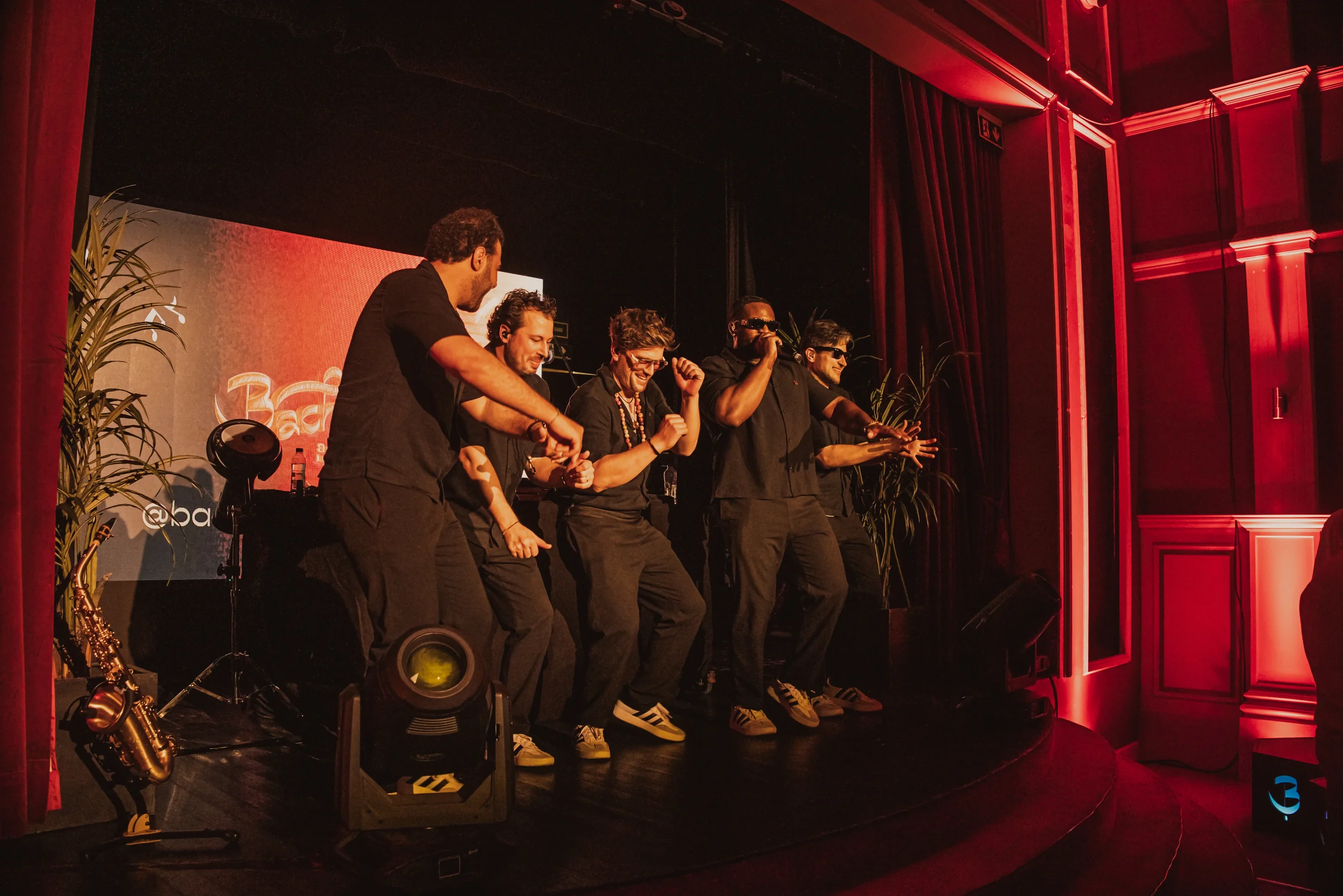 Joseph, Mike, Laurens, Angel en Loris. Vijf mannen die op een podium dansen en genieten, met een rode achtergrond en decoratie, en muziekequipment op de voorgrond.