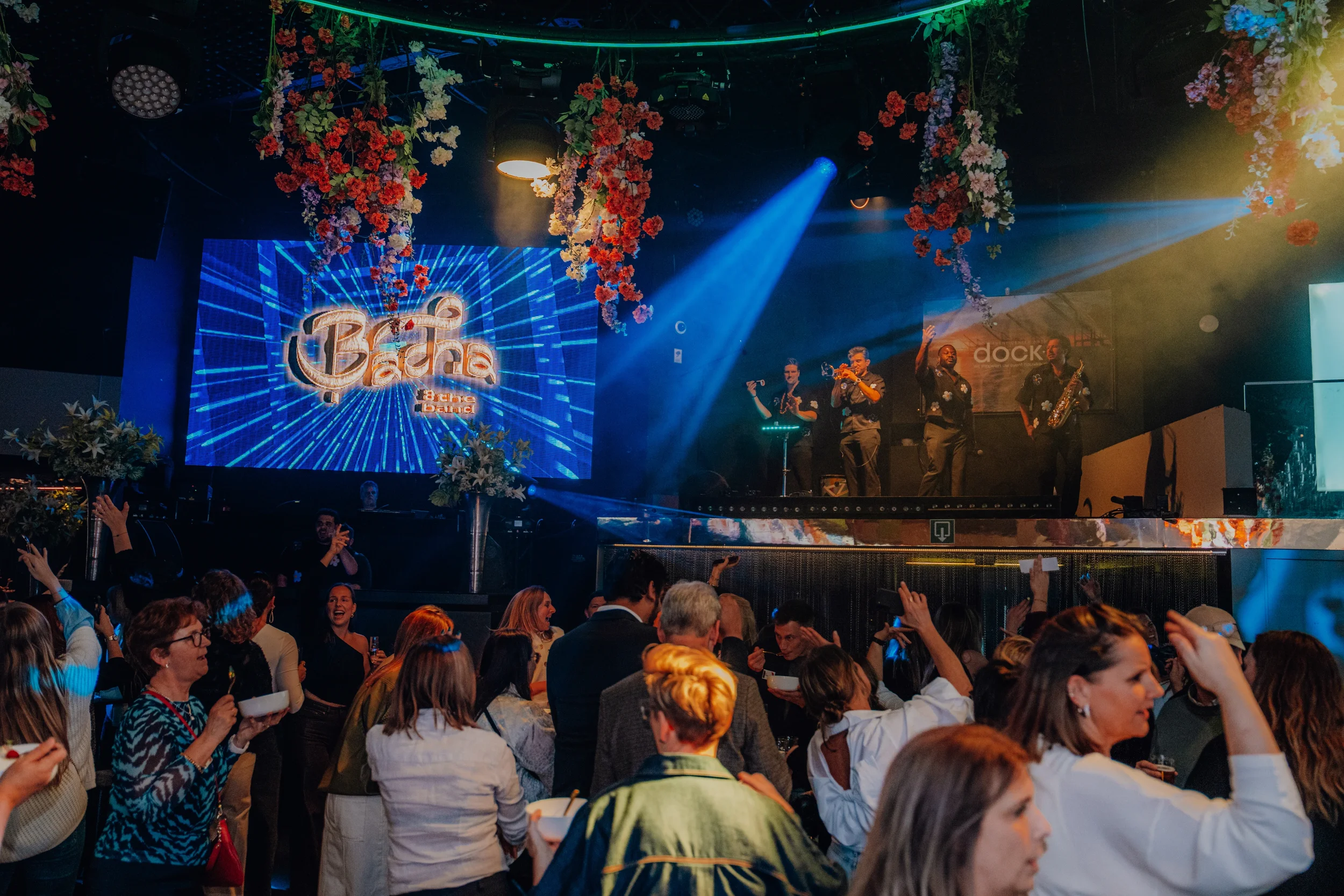 Bezoekers genieten van een live muziekoptreden van Bacha & The Band in een club, met een groot scherm op de achtergrond, versierd met kleurrijke bloemen en verlichting.
